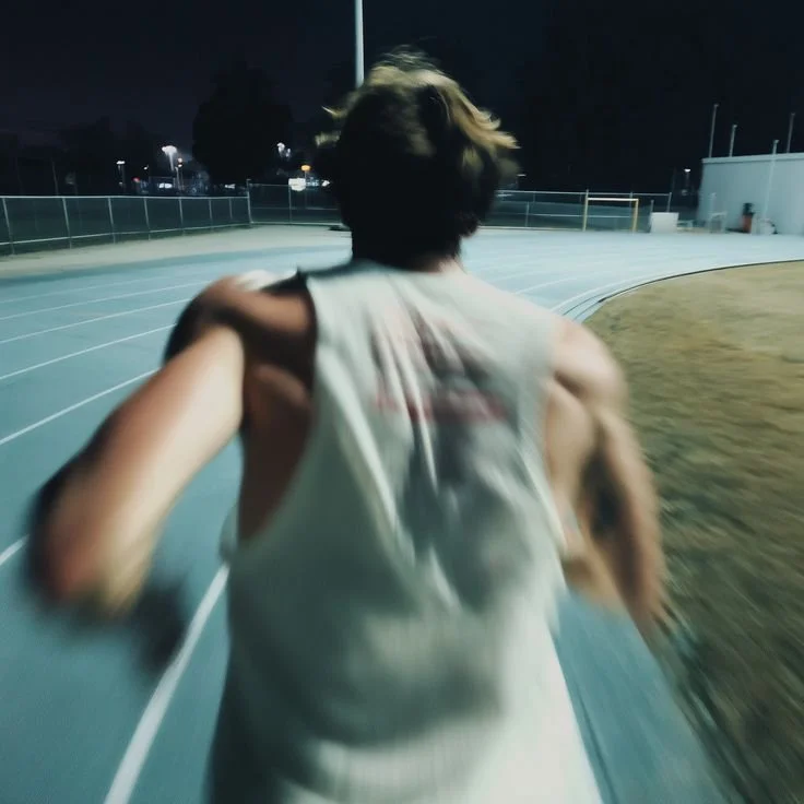 A person with shoulder-length hair running on a track at night, wearing a sleeveless shirt.