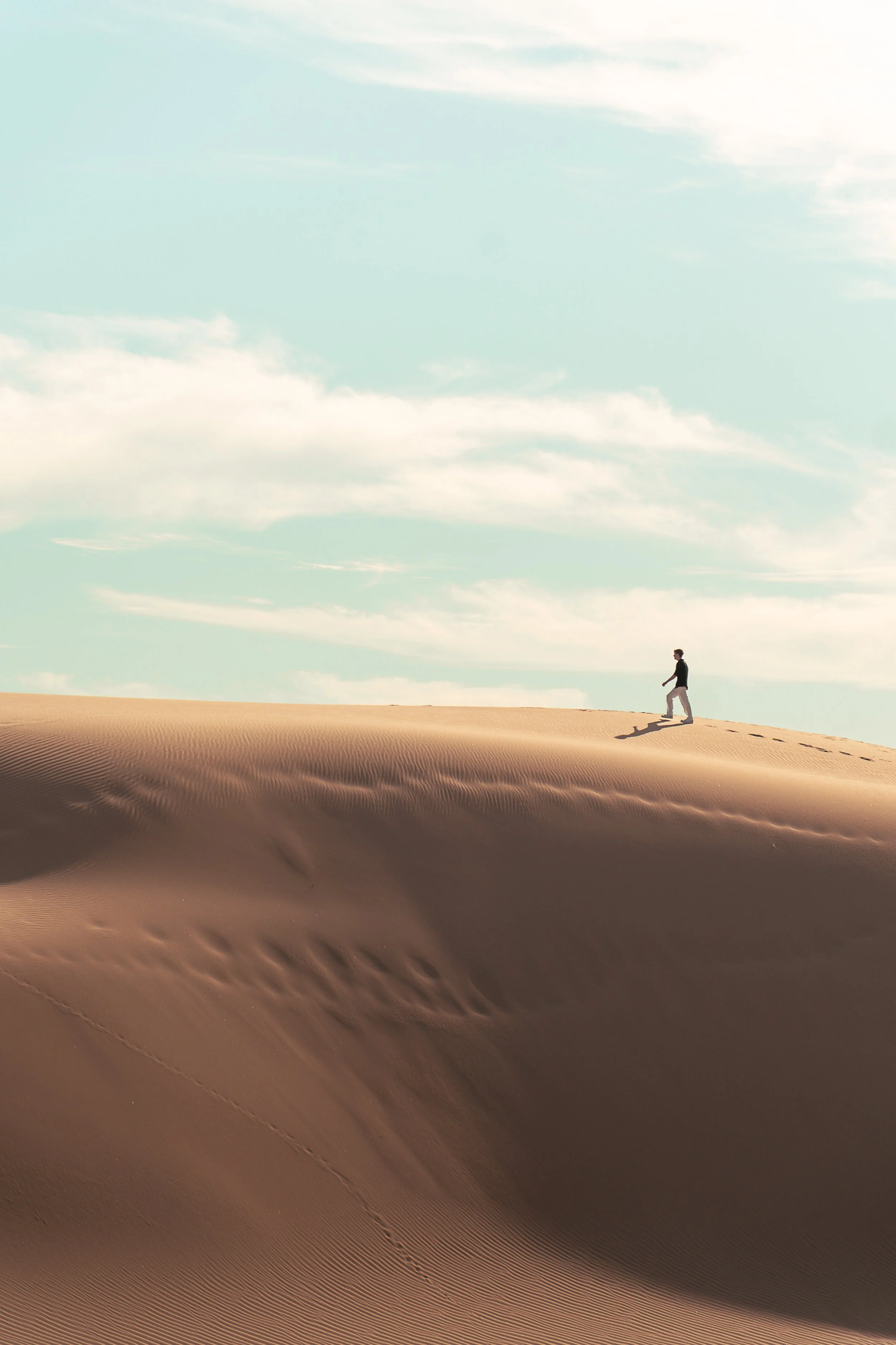 A person walking across sand dunes in a desert with a blue sky and scattered clouds overhead.