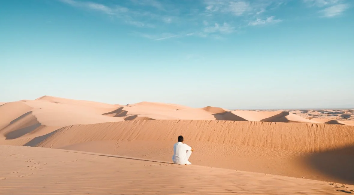 A man sitting alone on sand dunes in a desert under a blue sky.