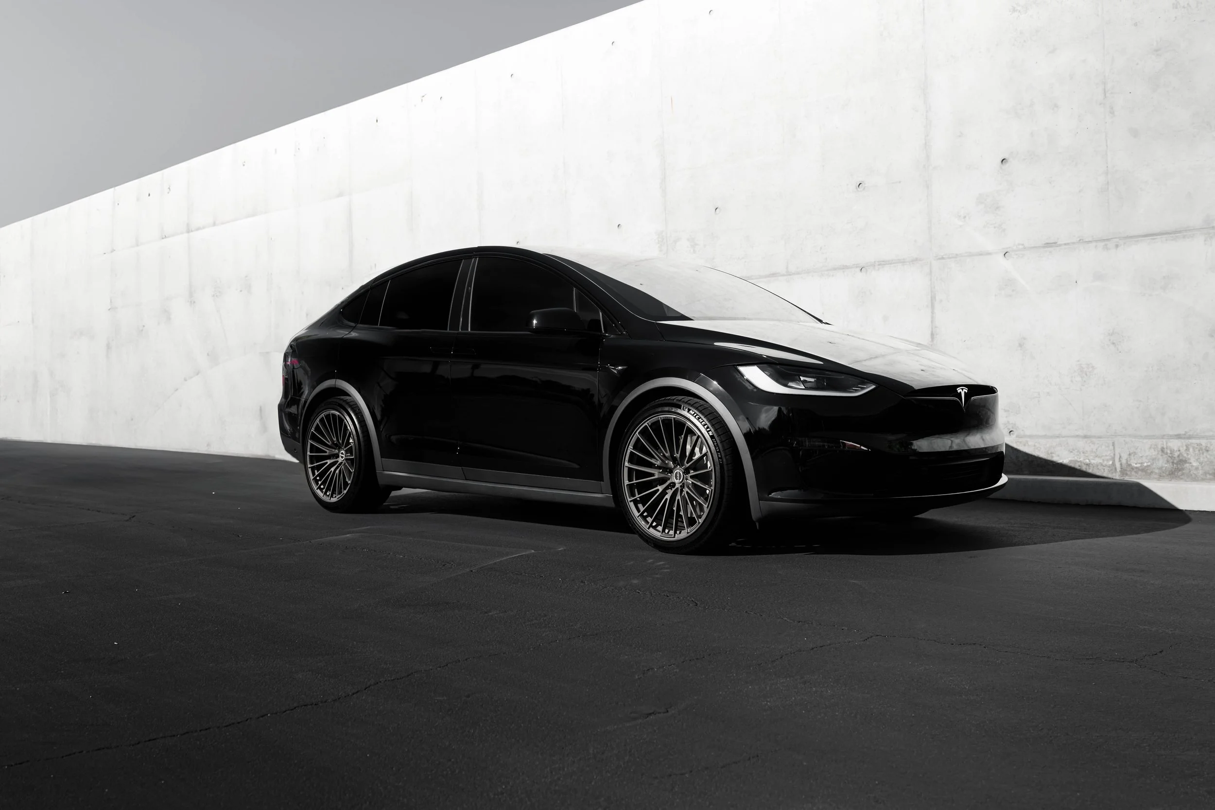 A black Tesla Model 3 electric car parked against a concrete wall in an outdoor setting, with the front of the car facing to the right.