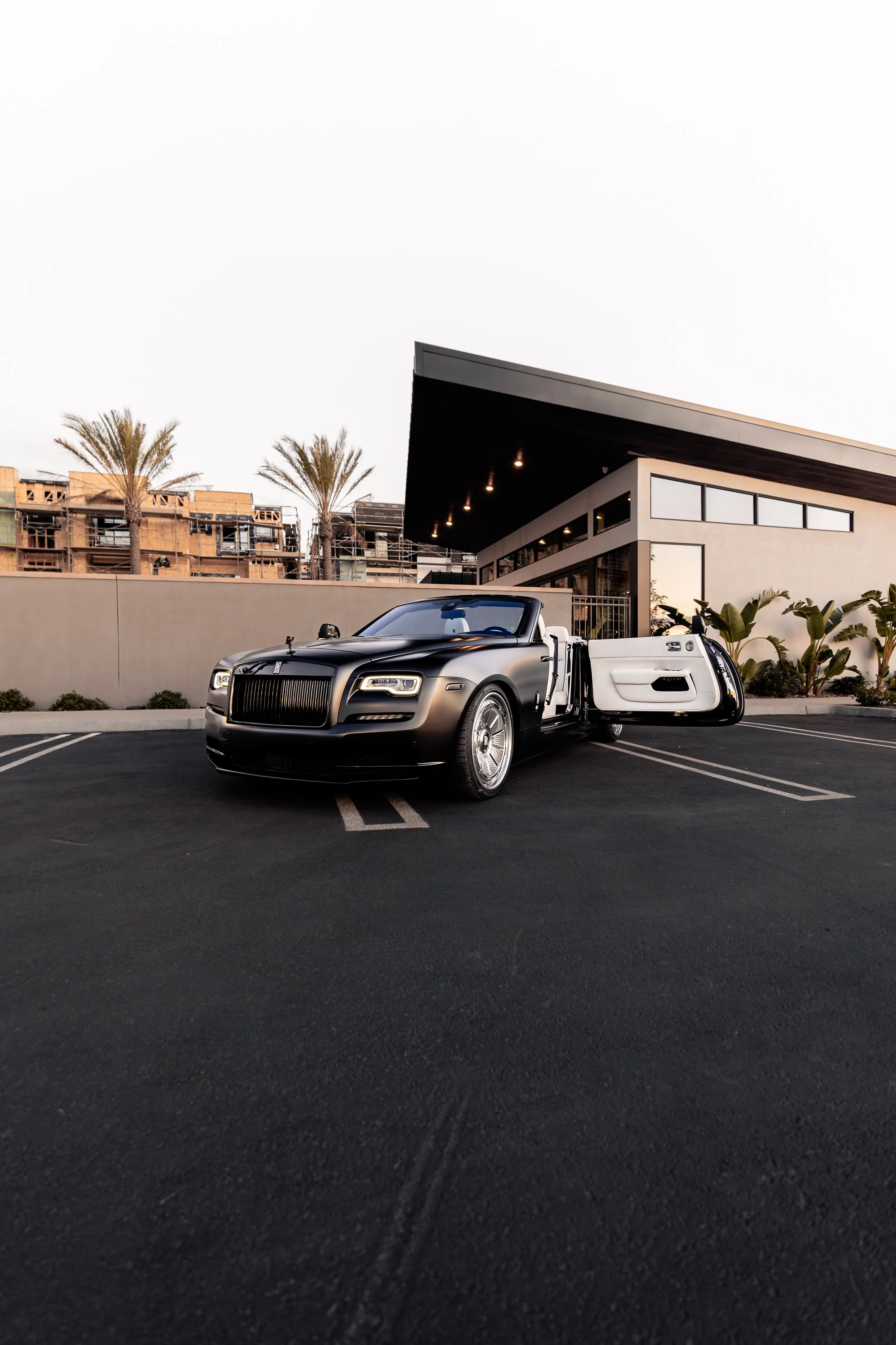 A black luxury convertible car with a white interior parked in an empty parking lot in front of a modern building with large windows and palm trees.