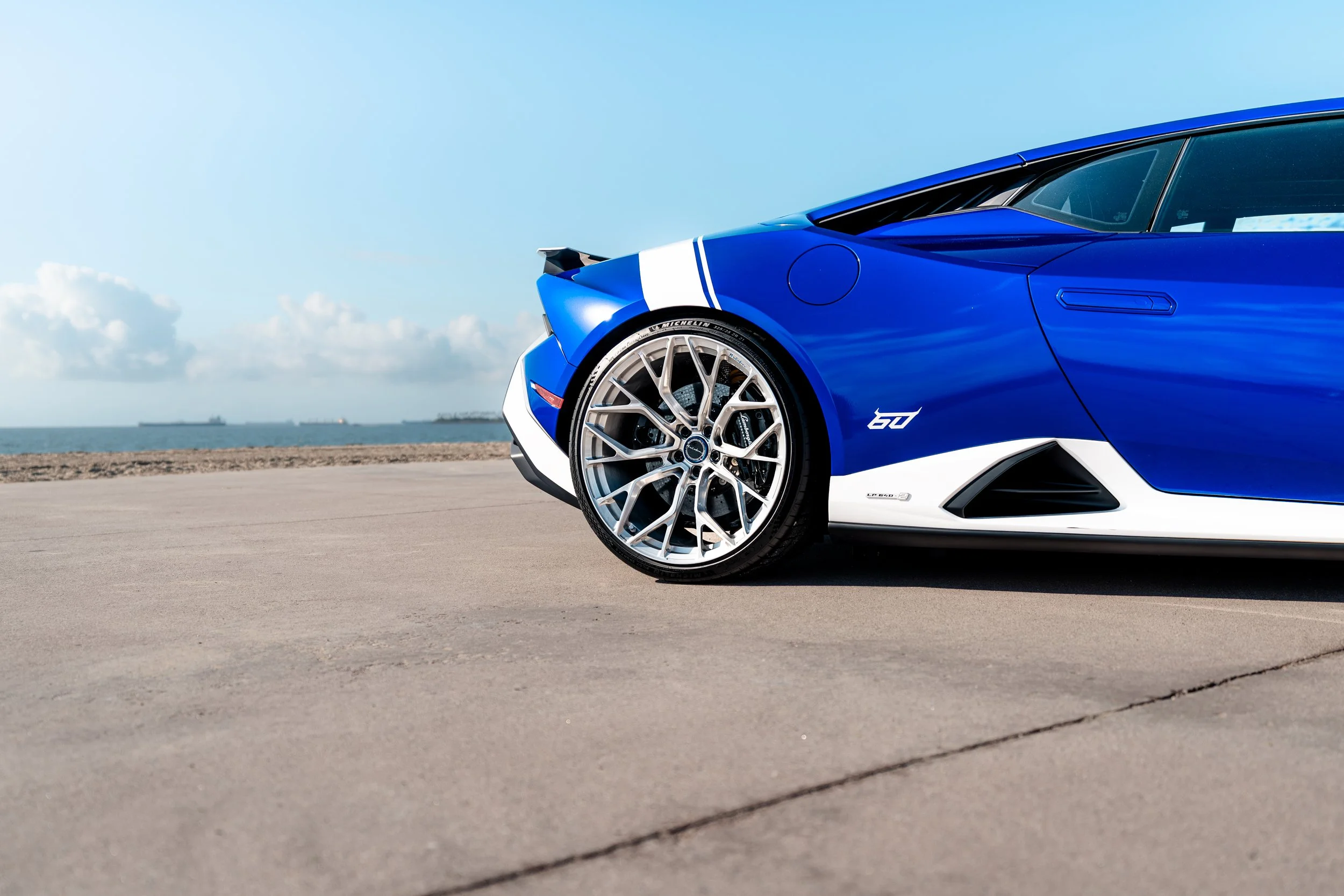 Close-up of a blue sports car with white accents parked on a coastal road, ocean in the background.