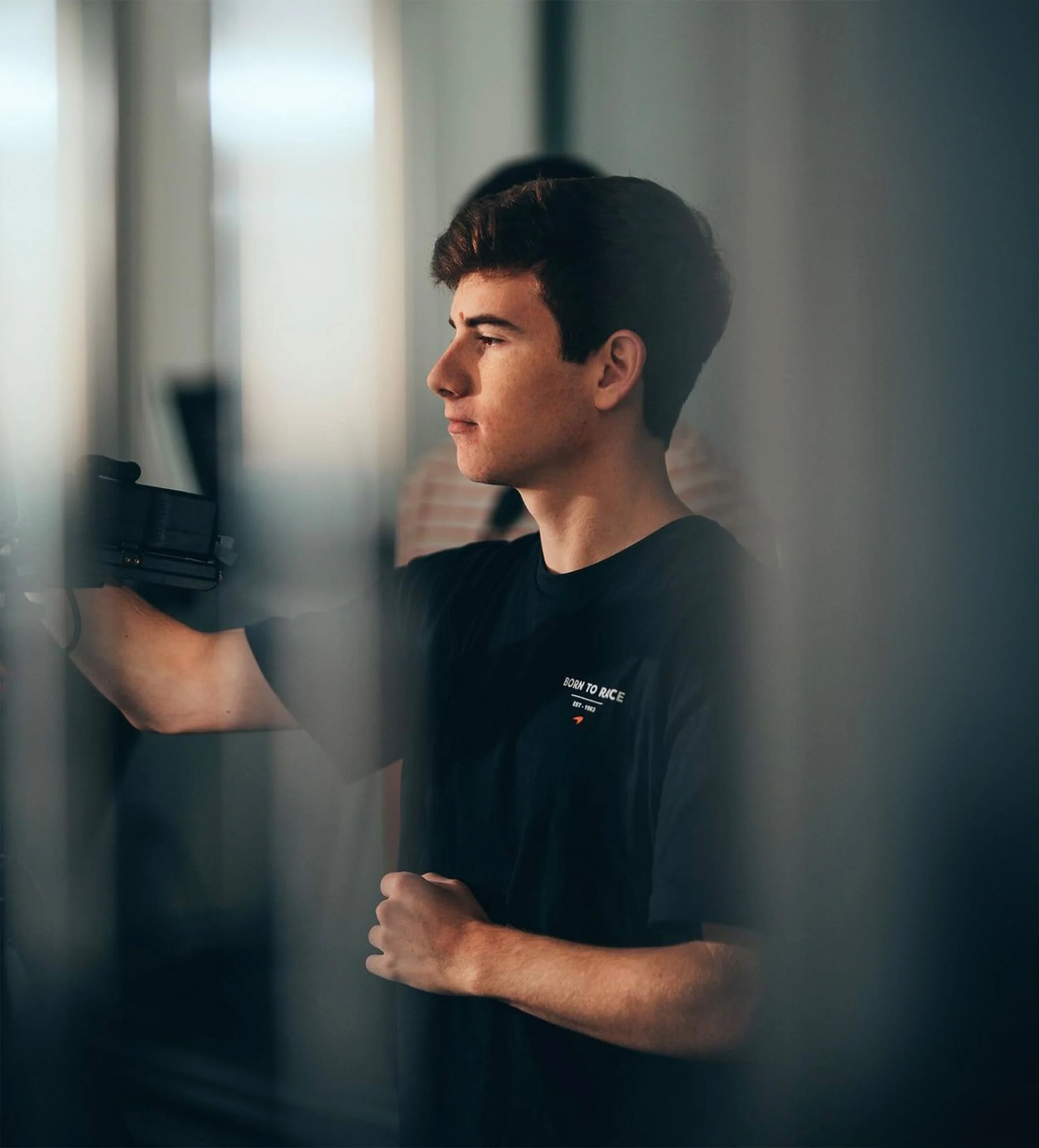 A young man with short dark hair and light skin, wearing a black T-shirt, is playing an arcade game or pinball machine. His expression is focused and he is holding a game controller or handle with his right hand, with his left fist clenched close to his chest. The background is blurry with other people and light coming from the left side.