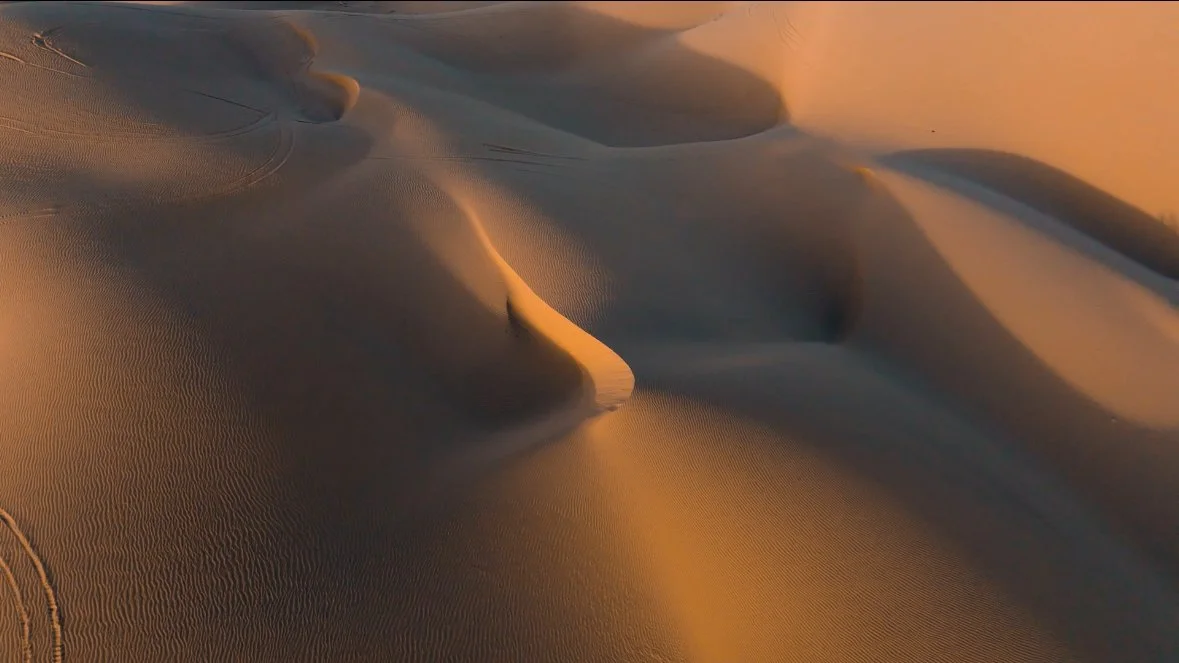 Sand dunes in a desert during sunset with shadows highlighting the ripples and textures in the sand.