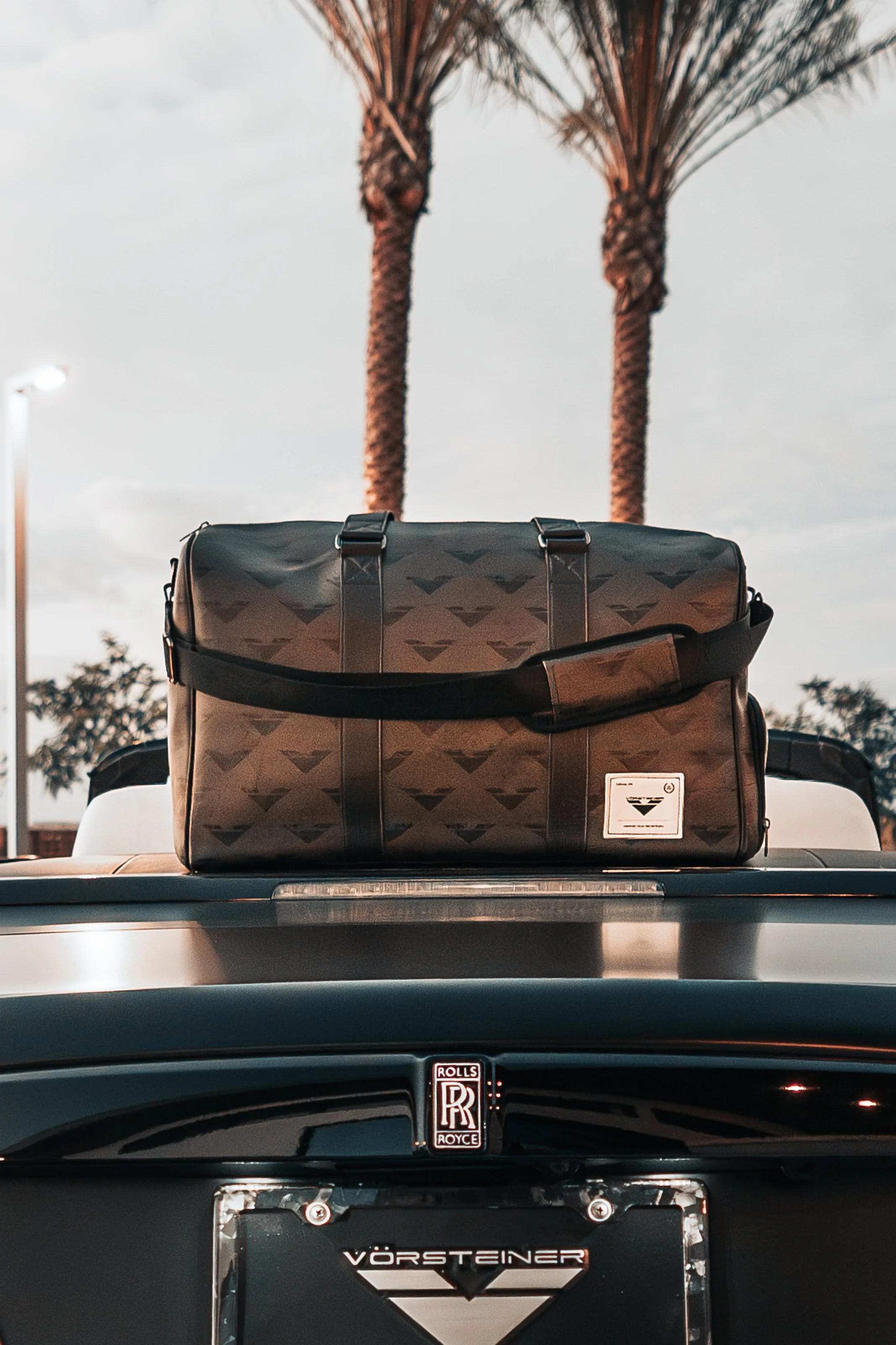 A black collar bag with a geometric pattern resting on the rear of a black Rolls Royce car with Palm trees in the background at sunset.