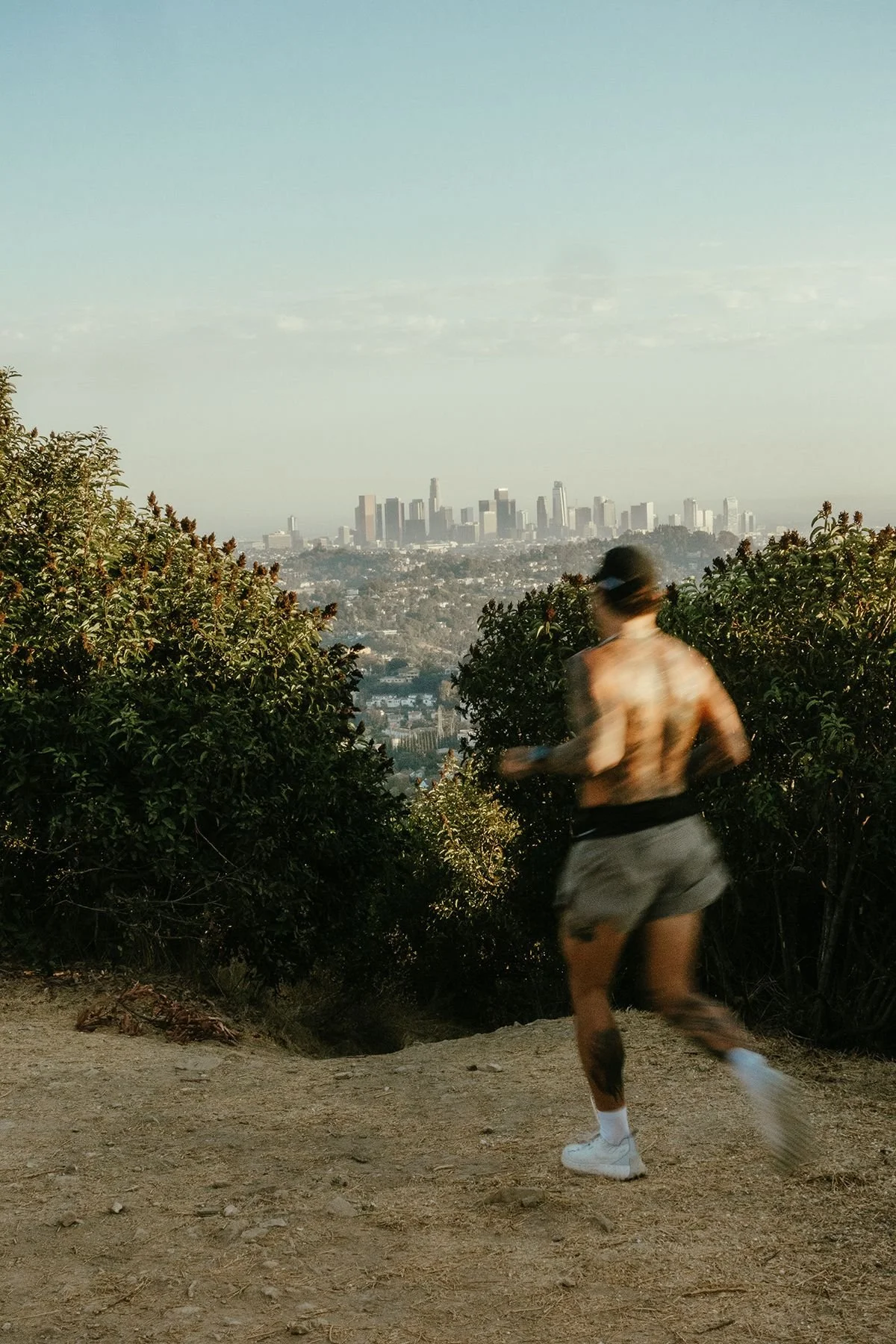 A person jogging on a trail with a city skyline in the distance, lush foliage on either side, and a partly cloudy sky overhead.