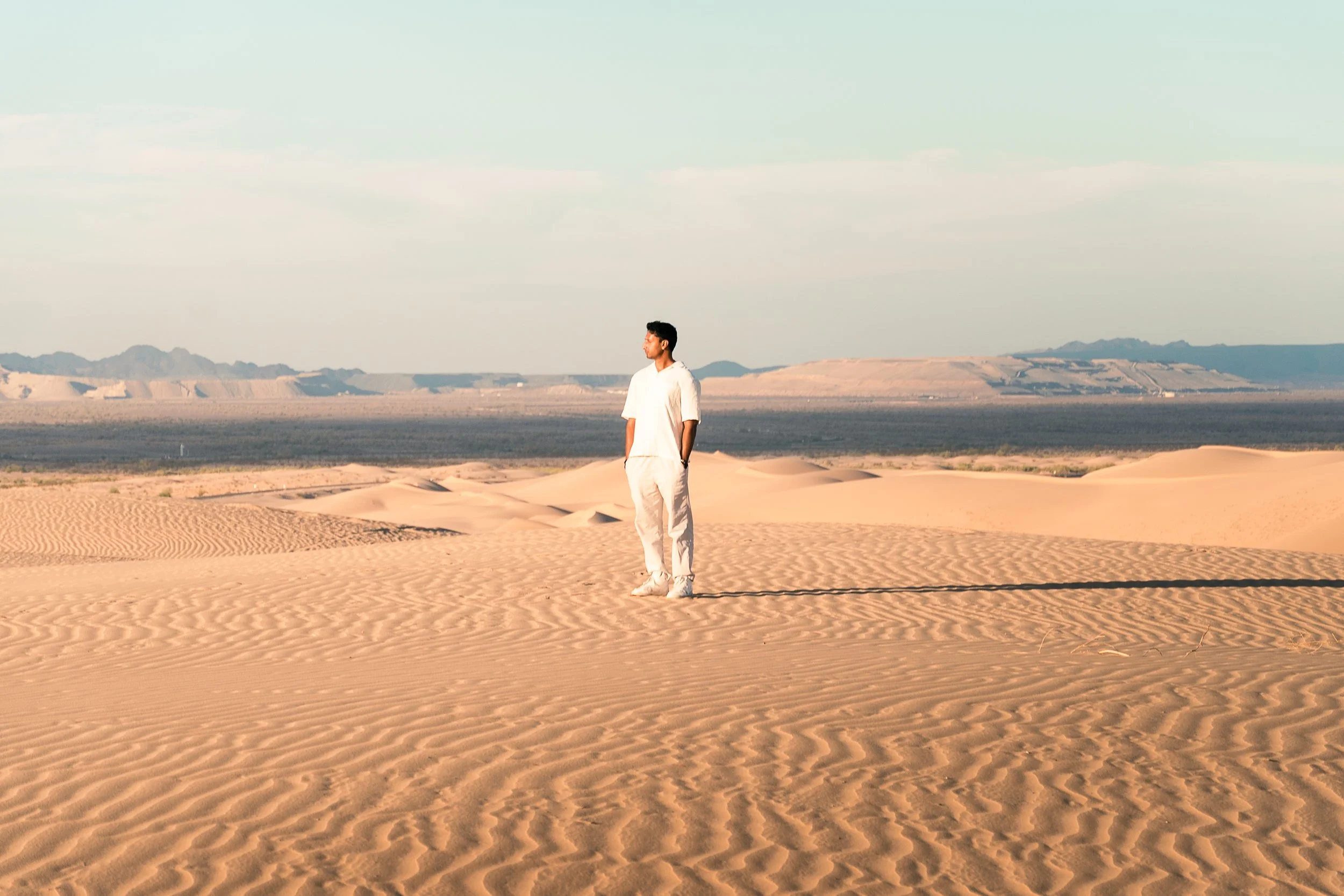 A man wearing white clothing standing alone in a desert, with sand dunes and mountains in the background under a clear sky.