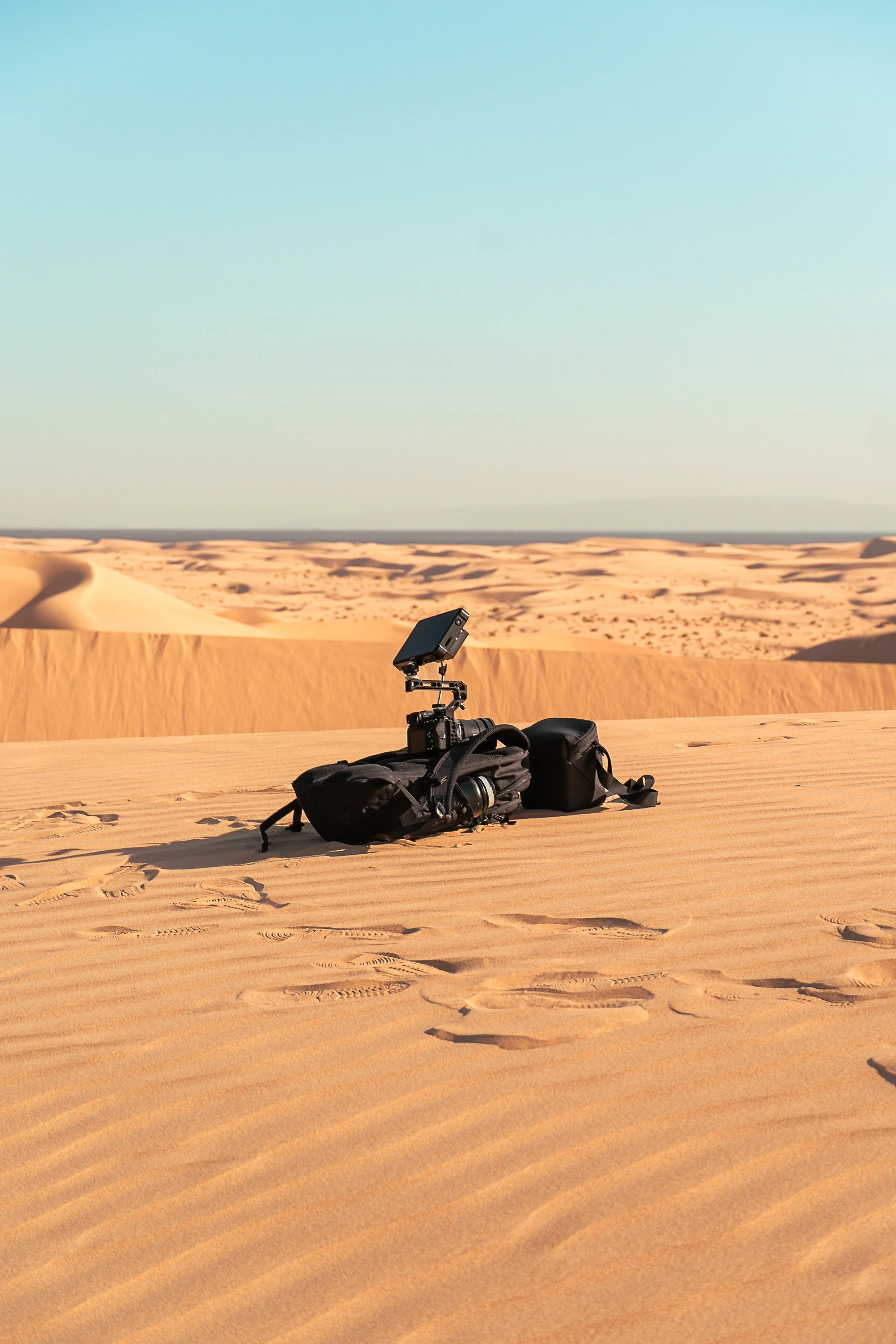 A camera and backpack placed on the sand of a desert with sand dunes in the background under a clear sky.