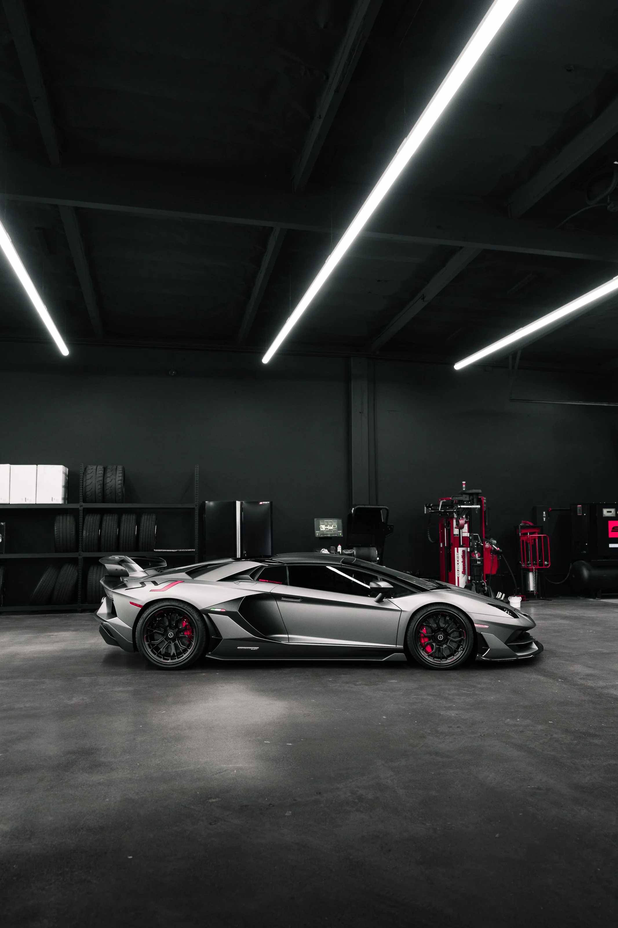 Black and silver Lamborghini sports car inside a garage with shelves of tires and equipment in the background.