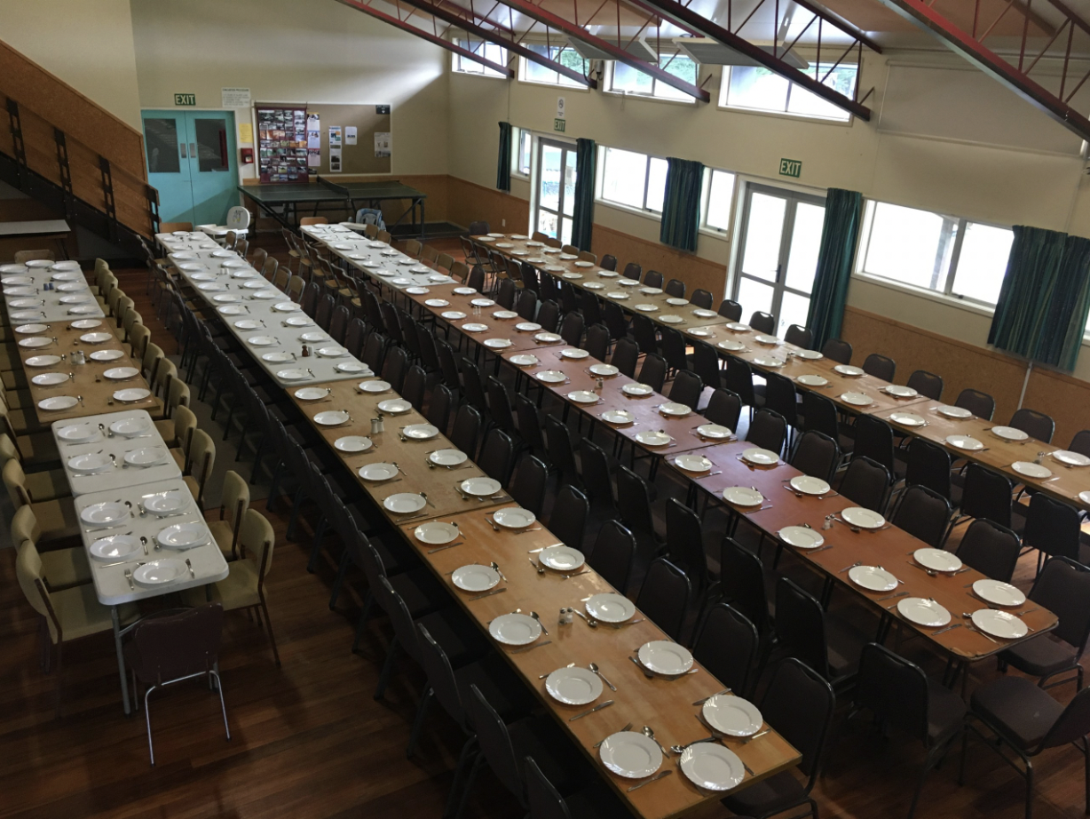Large dining hall with long tables set with white plates, forks, knives, and napkins, prepared for a meal, with several chairs around each table, and windows along the wall with green curtains.