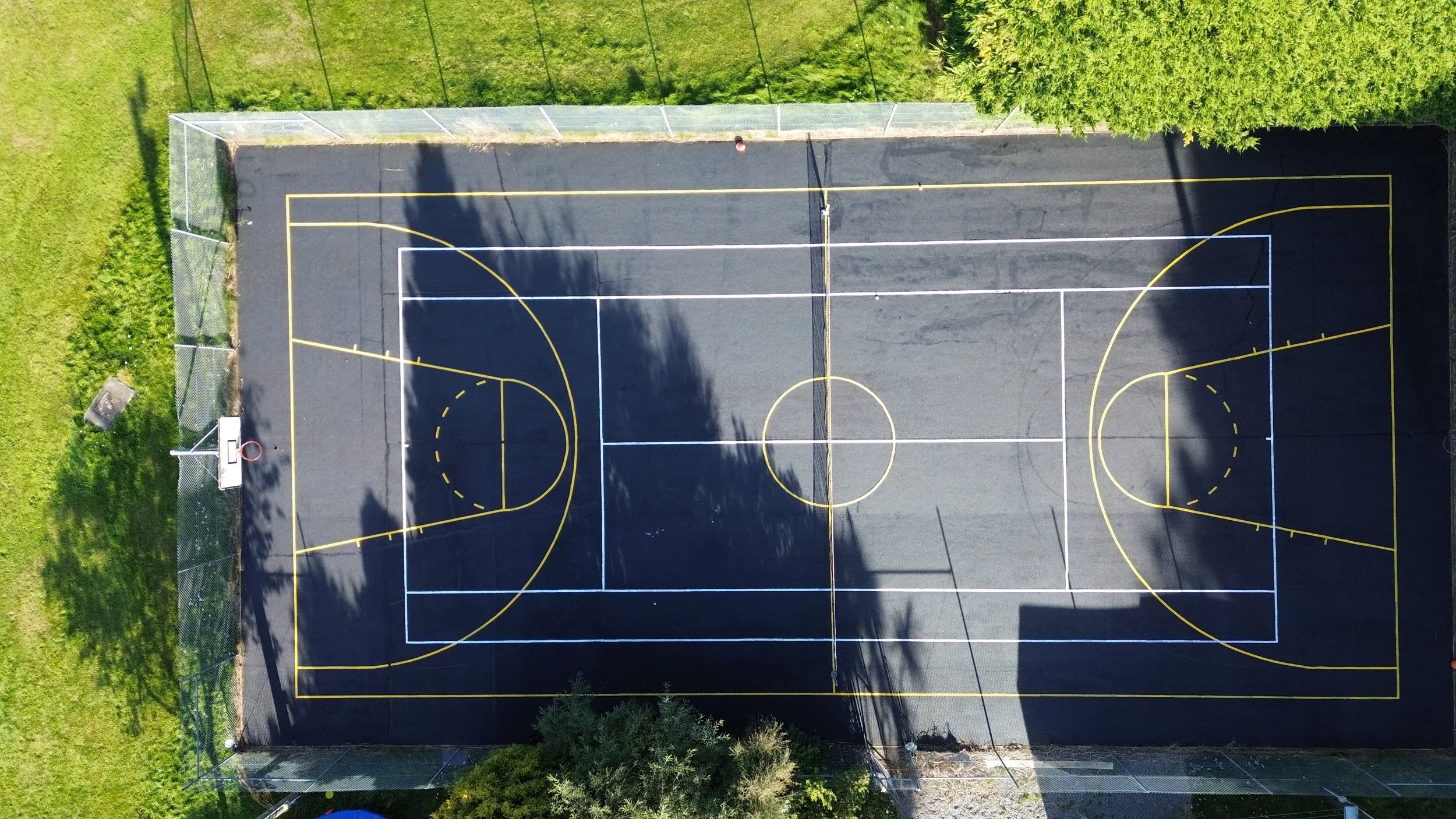 An aerial view of a basketball court with white and yellow lines marking the playing area, surrounded by green grass and trees, with a basketball hoop in the lower left corner.