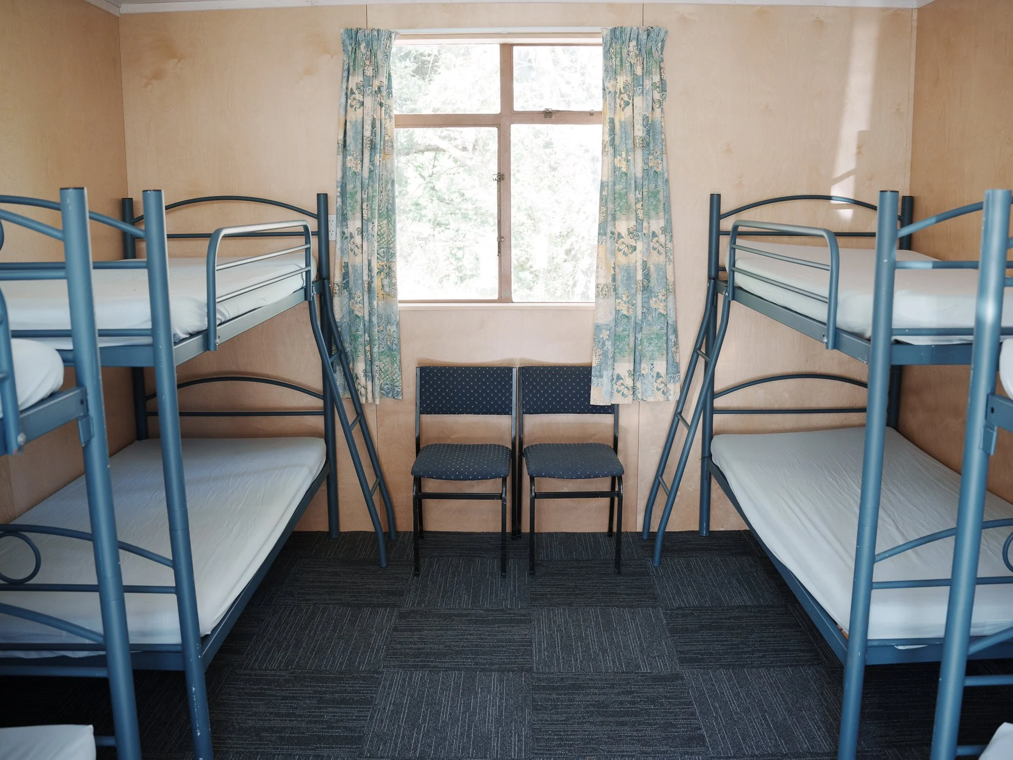 A simple dorm room with four metal bunk beds, two on each side, and two chairs in the center facing each other. A window with floral curtains is in the middle of the back wall, letting in natural light.