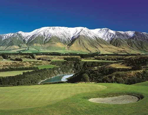 A golf course with a sand trap, lush green fairways, a winding river, and snow-capped mountains in the background under a clear blue sky.
