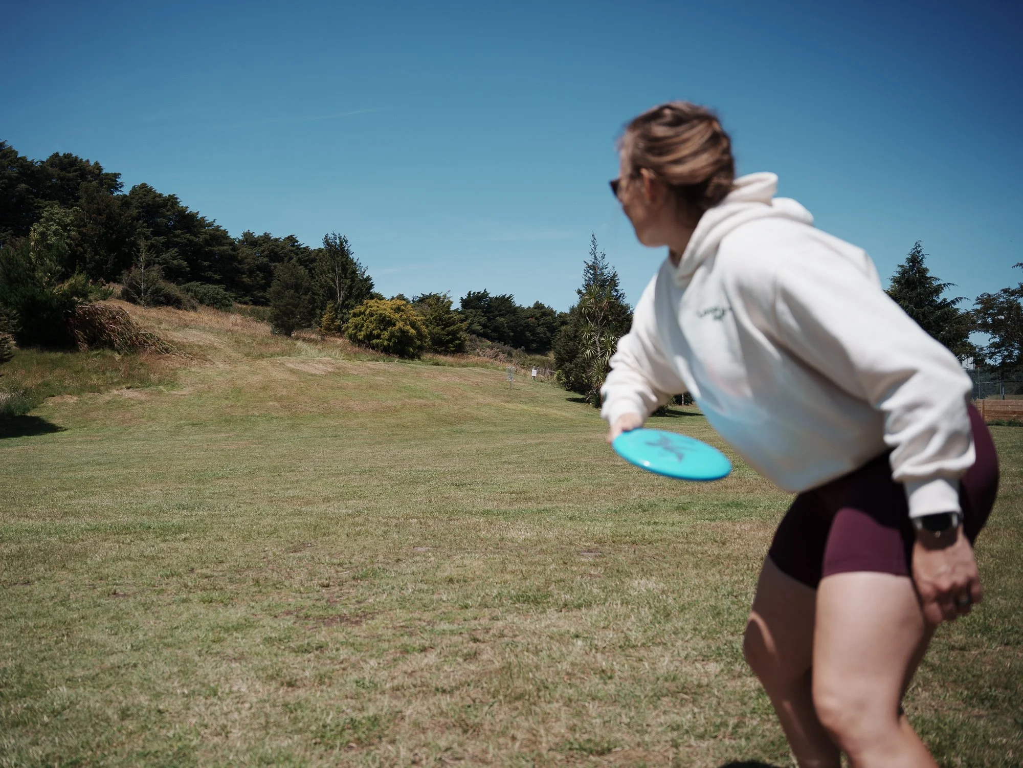 A woman in athletic attire playing disc golf outdoors on a grassy field with trees in the background under a clear blue sky.