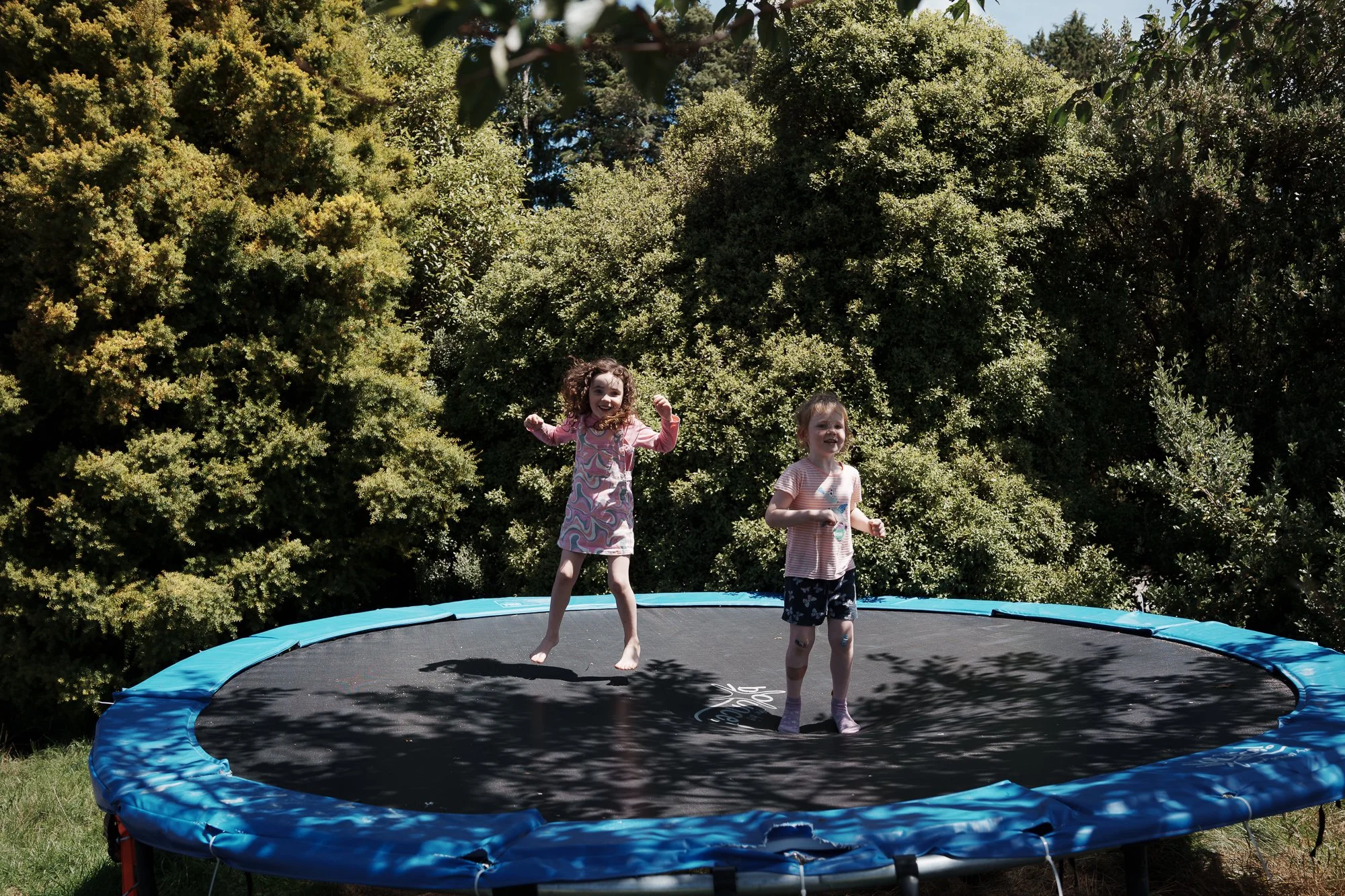 Two young girls are jumping and playing on a trampoline outdoors surrounded by trees on a sunny day.