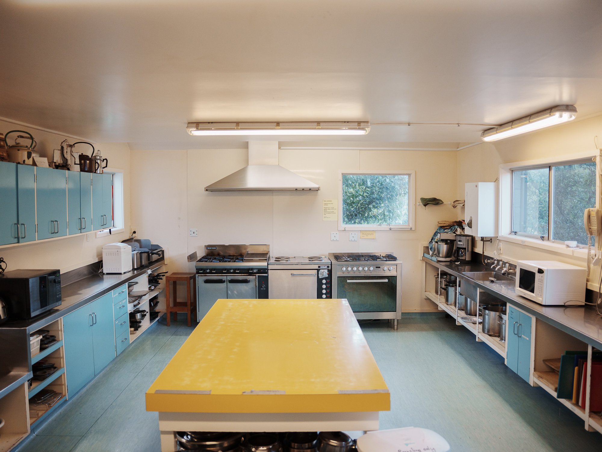 A spacious kitchen with light yellow walls, blue cabinets, and large windows. Contains cooking appliances like stoves, microwave, and kitchen tools, with a yellow table in the center.