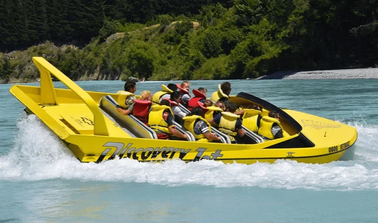 A yellow Discovery jet boat with several passengers wearing life jackets, speeding across a river with a wooded landscape in the background.