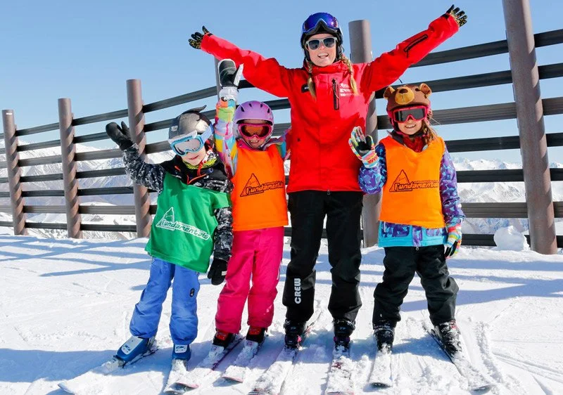 A group of four people, including children and an adult, on skis in a snowy mountain area, all wearing helmets and ski gear. They are smiling and raising their arms, with a wooden fence and snow-covered mountains in the background.