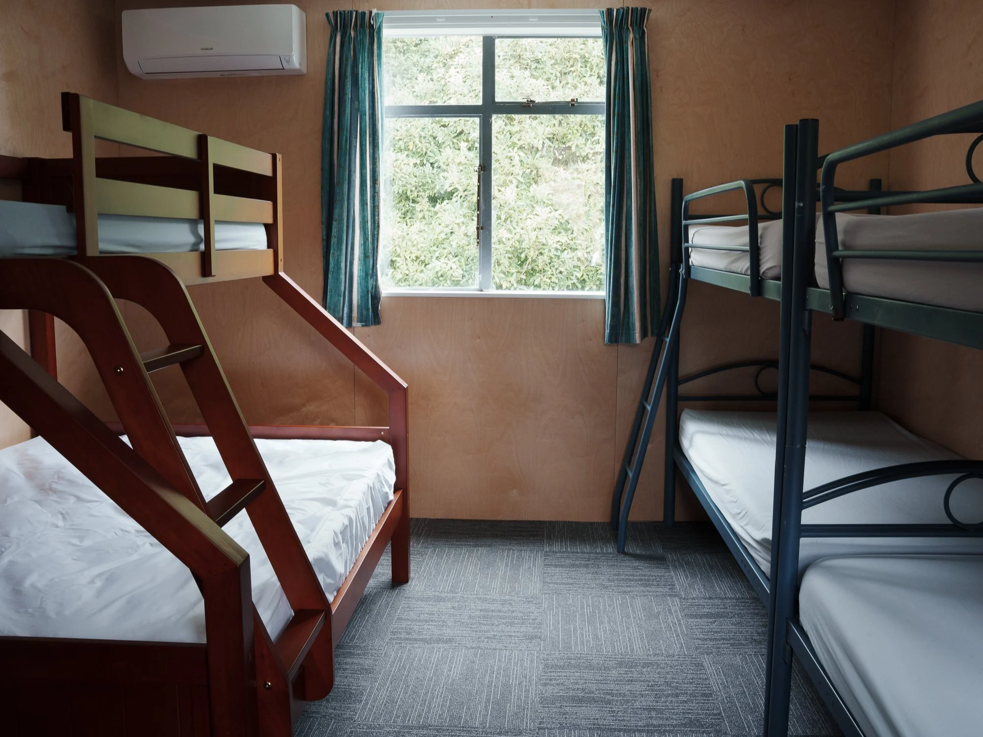 A small room with wooden walls containing three bunk beds, one made of wood and two of metal, with a window showing greenery outside, and a wall-mounted air conditioner.