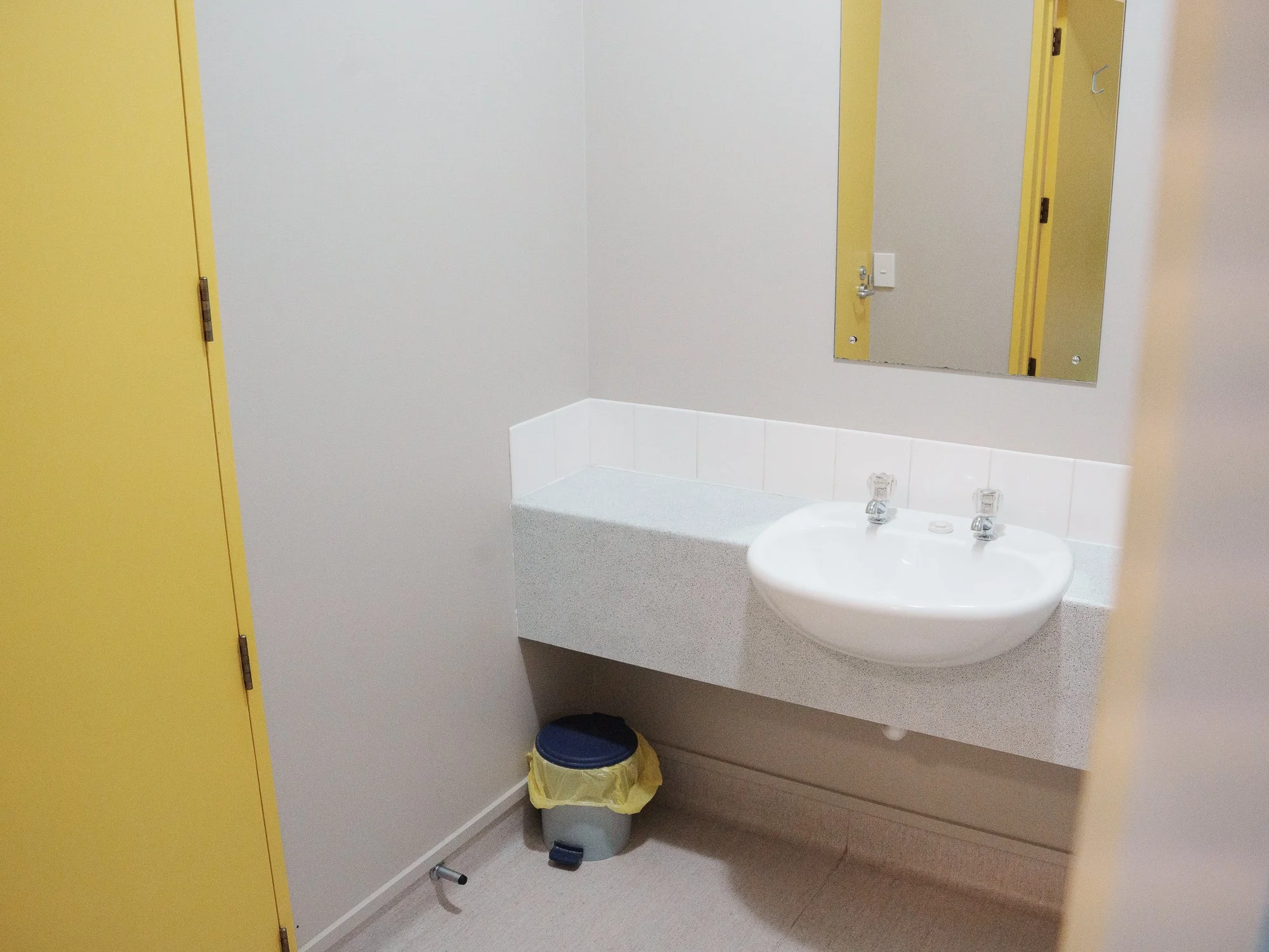 Empty bathroom with yellow door, mirror, white sink with two faucets, in a room with gray walls and beige floor.