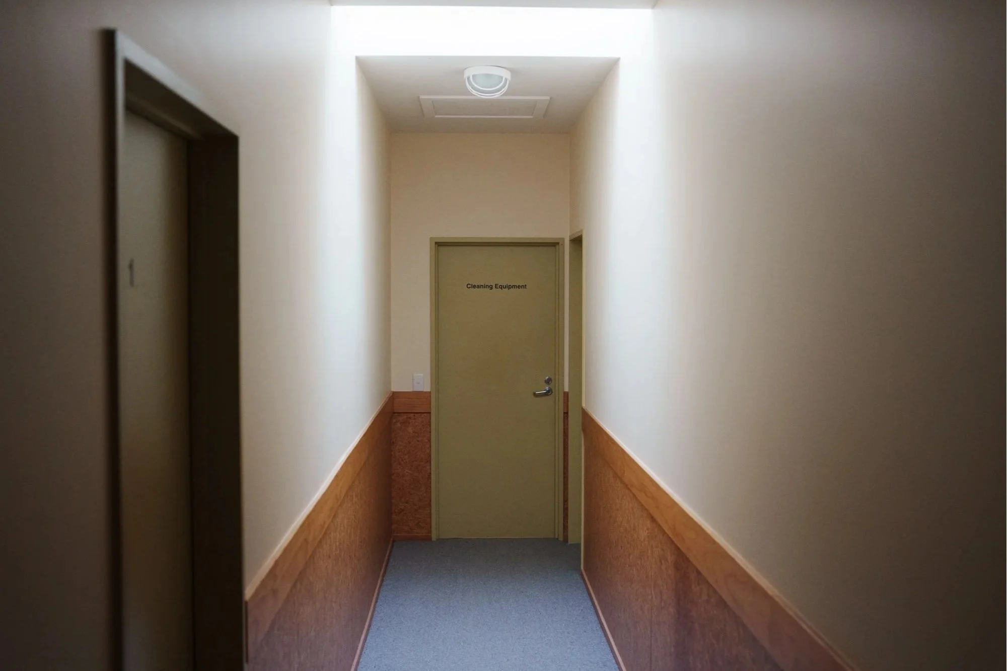A narrow corridor with beige walls, wood paneling, and a gray carpet. An elevator door is on the left, and a door labeled 'Cleaning Equipment' is at the end of the hallway.