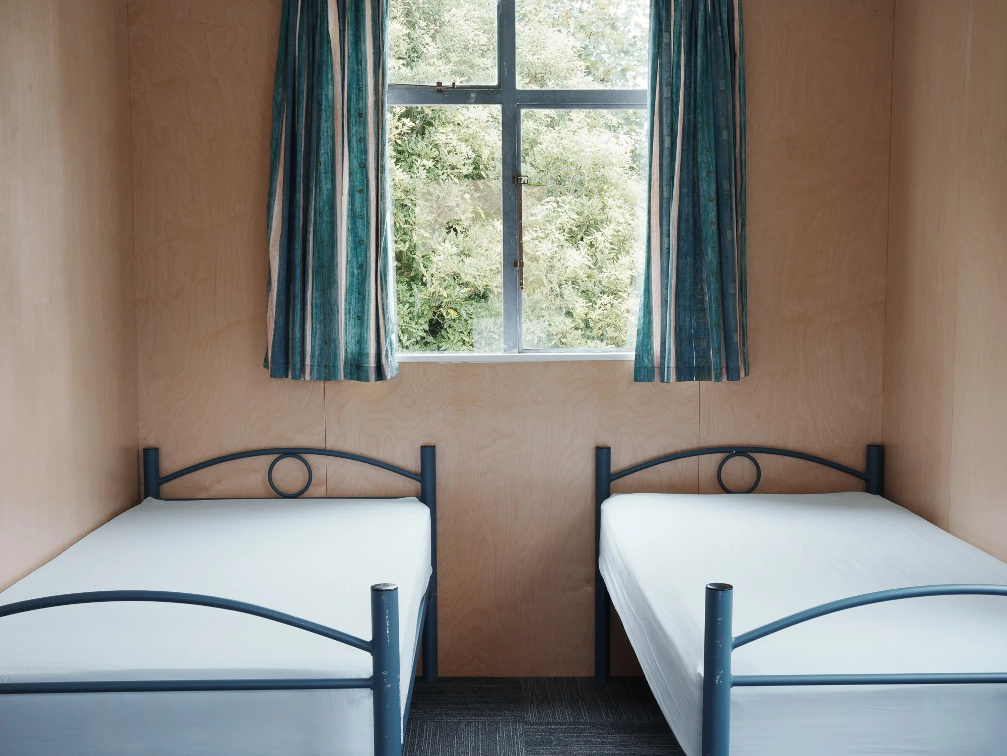 A simple bedroom with two single beds with white mattresses, dark metal frames, and a window with blue striped curtains that looks out to green foliage.