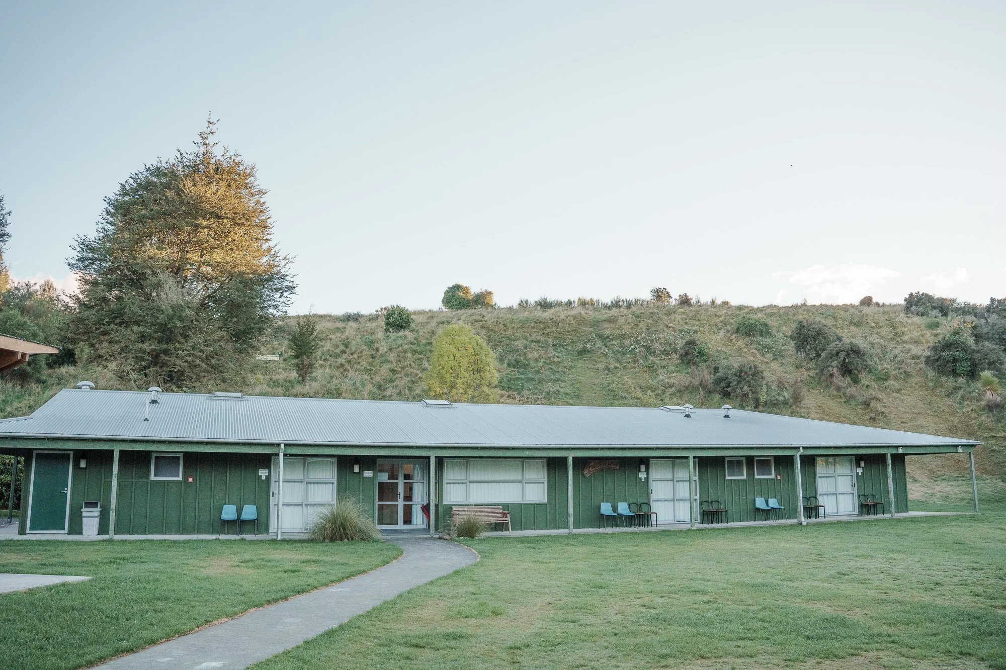 A single-story green building with a metal roof and outdoor seating, set against a grassy lawn and hillside with trees under a clear sky.