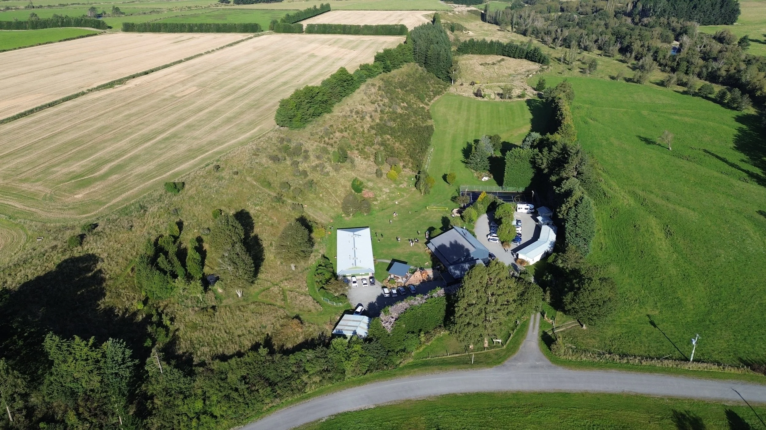 Aerial view of a rural property with several buildings, a parking lot, and surrounding green fields and trees.