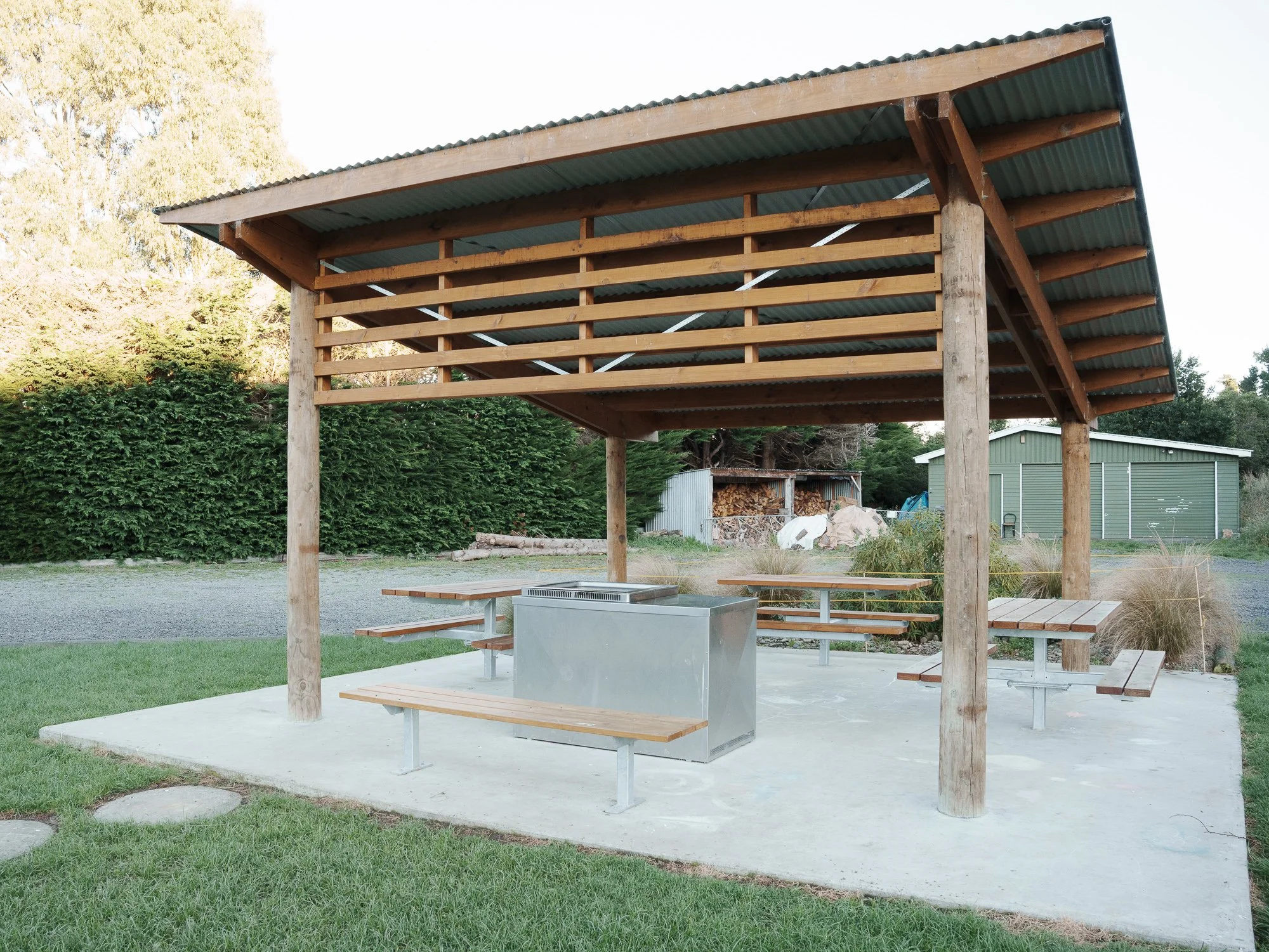 Outdoor shelter with a sloped roof, wooden railings, and multiple benches around a central stainless steel barbecue grill, situated on a concrete pad in a grassy area with trees and a small building in the background.