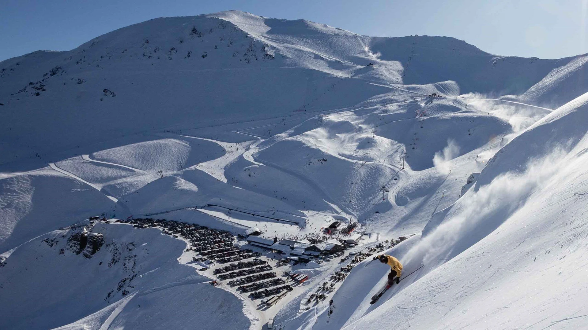 A snowy mountain with ski slopes, ski lifts, and a parking lot below. A skier is descending the slope in the foreground.