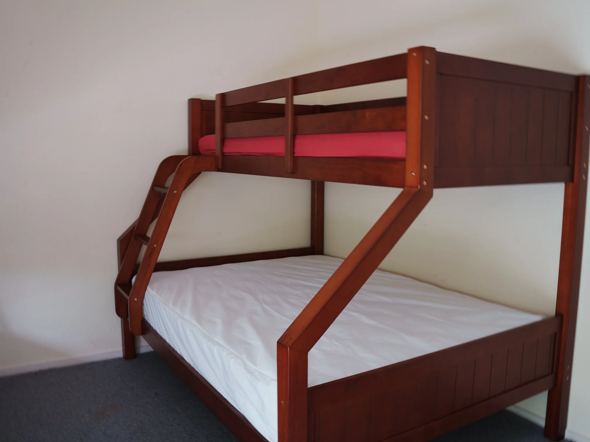 Wood bunk bed with a ladder, a pink mattress on the top bunk, and a white mattress on the bottom bunk, set against a plain white wall.