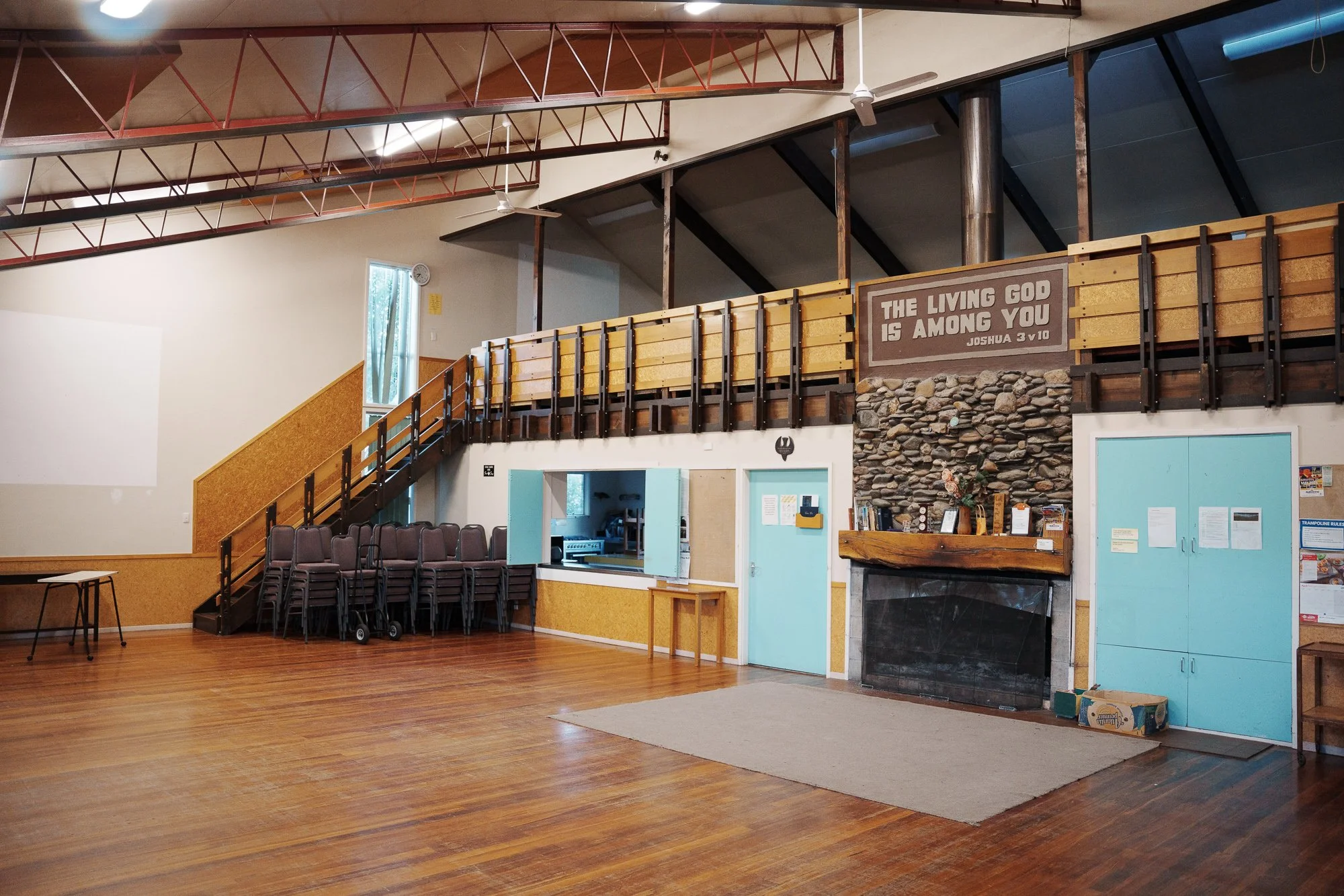 Empty community hall with a stack of chairs, a small table, a fireplace, and a sign that reads, "The living God is among you, Joshua 3 v 10."
