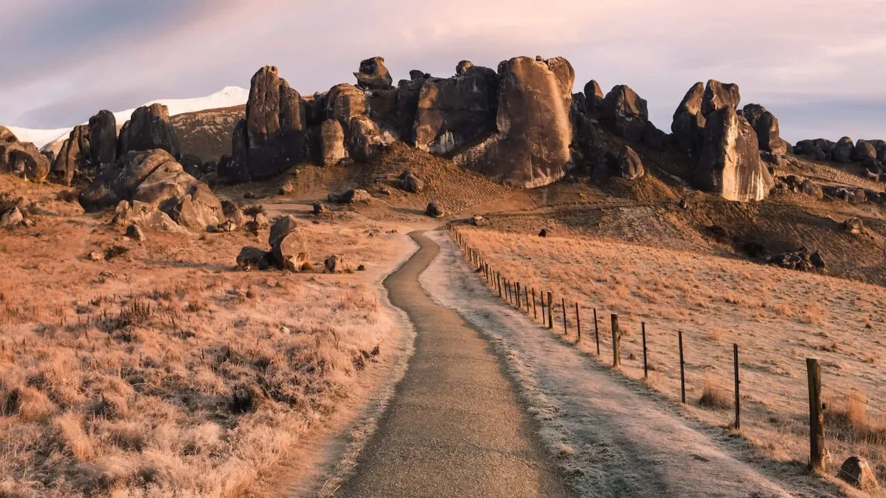 A winding dirt path through a dry, grassy field leading towards large rocky formations on a hill in the distance, with a fence on one side and snow-capped mountains under a pinkish sky.