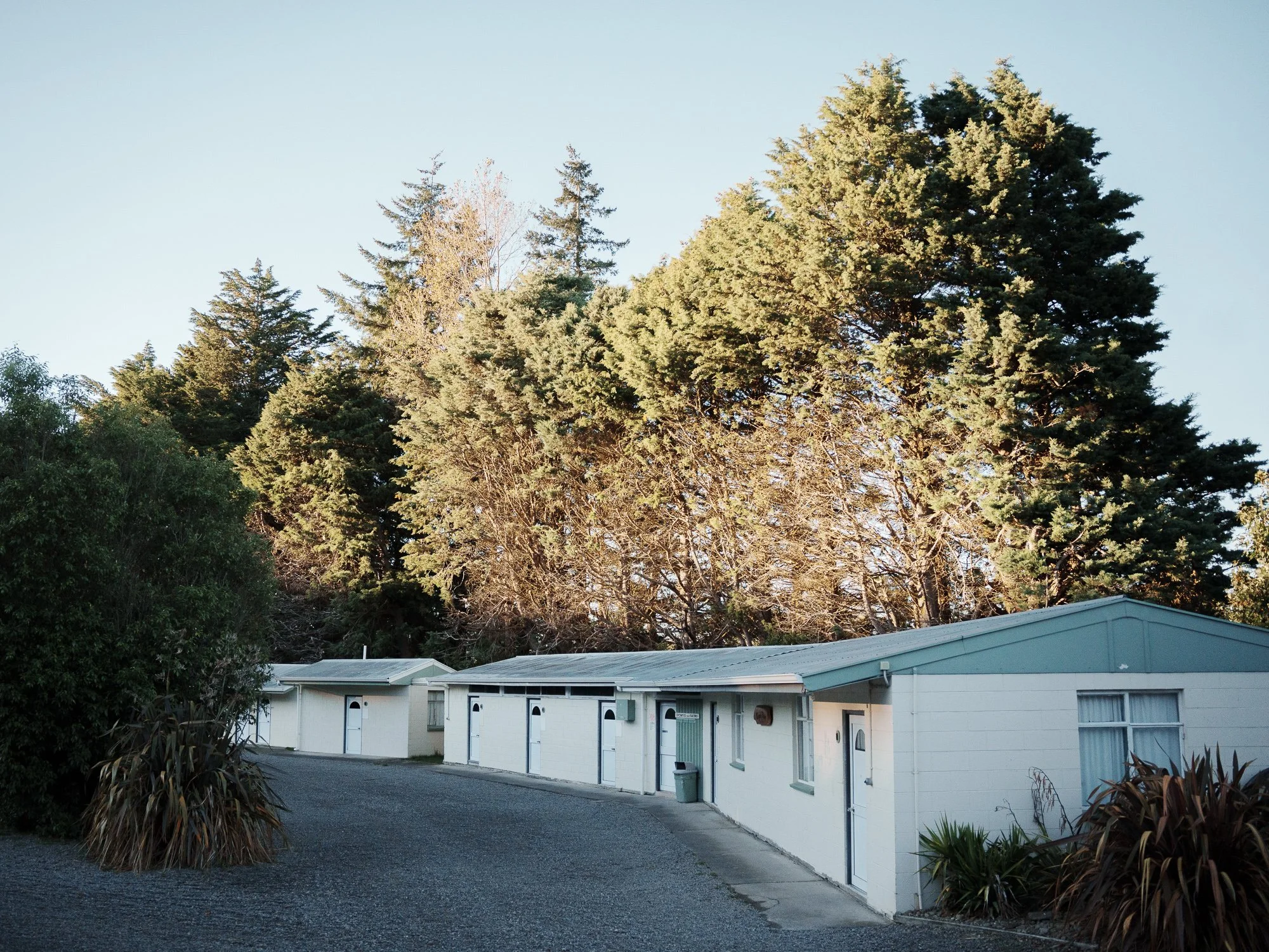 A row of white cabins with closed garage doors, set against a backdrop of tall, green trees under a clear sky.
