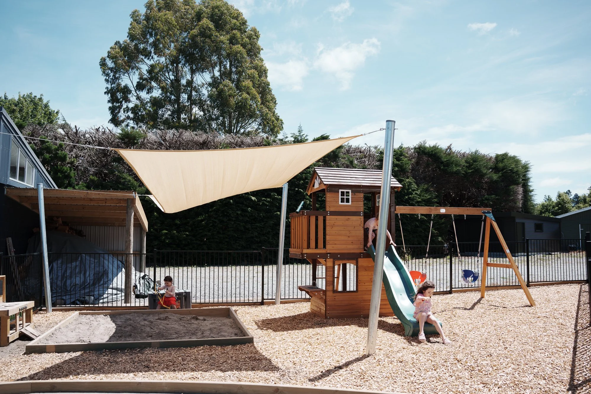 Children playing on a wooden playground in a sandbox and sliding on a slide, with swings and a shade sail, enclosed by a black fence, with trees and a blue sky in the background.