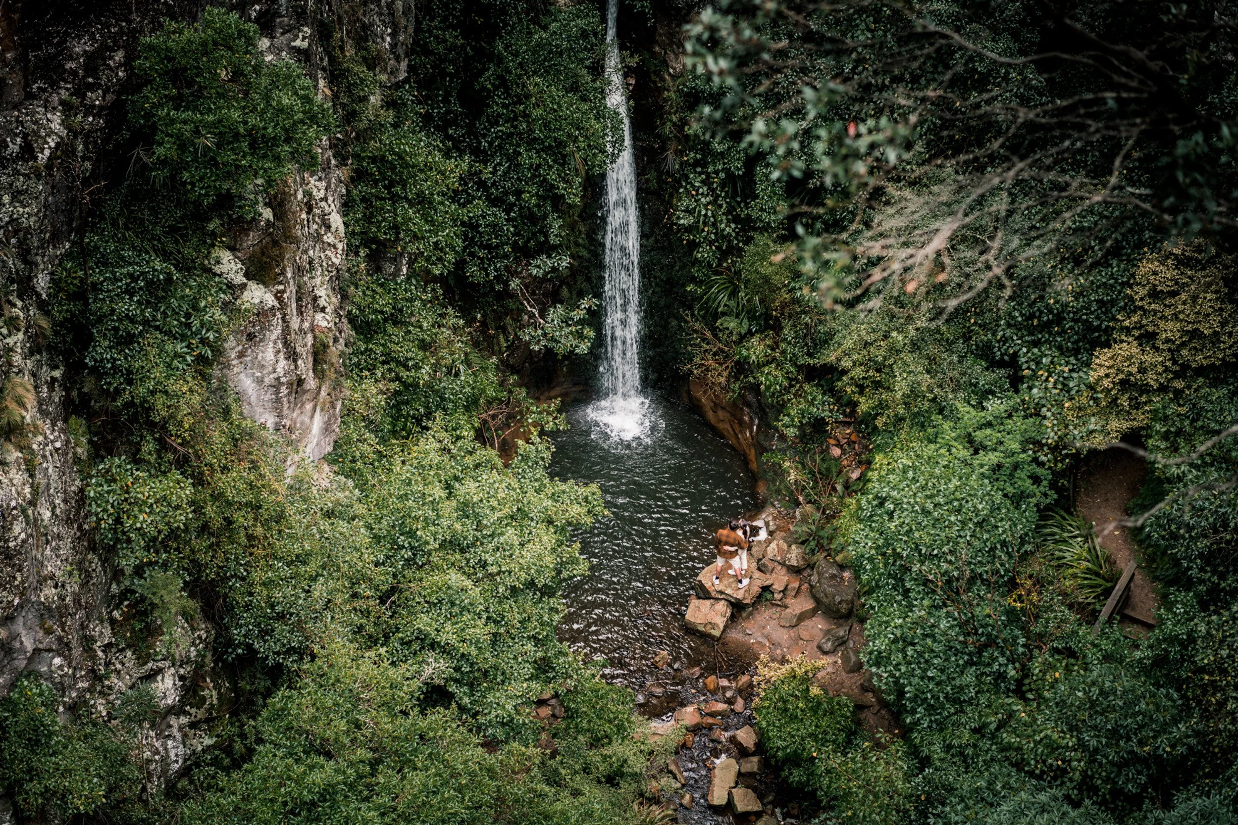 A waterfall cascading into a small pool surrounded by dense green forest foliage, with a person sitting on rocks near the water.