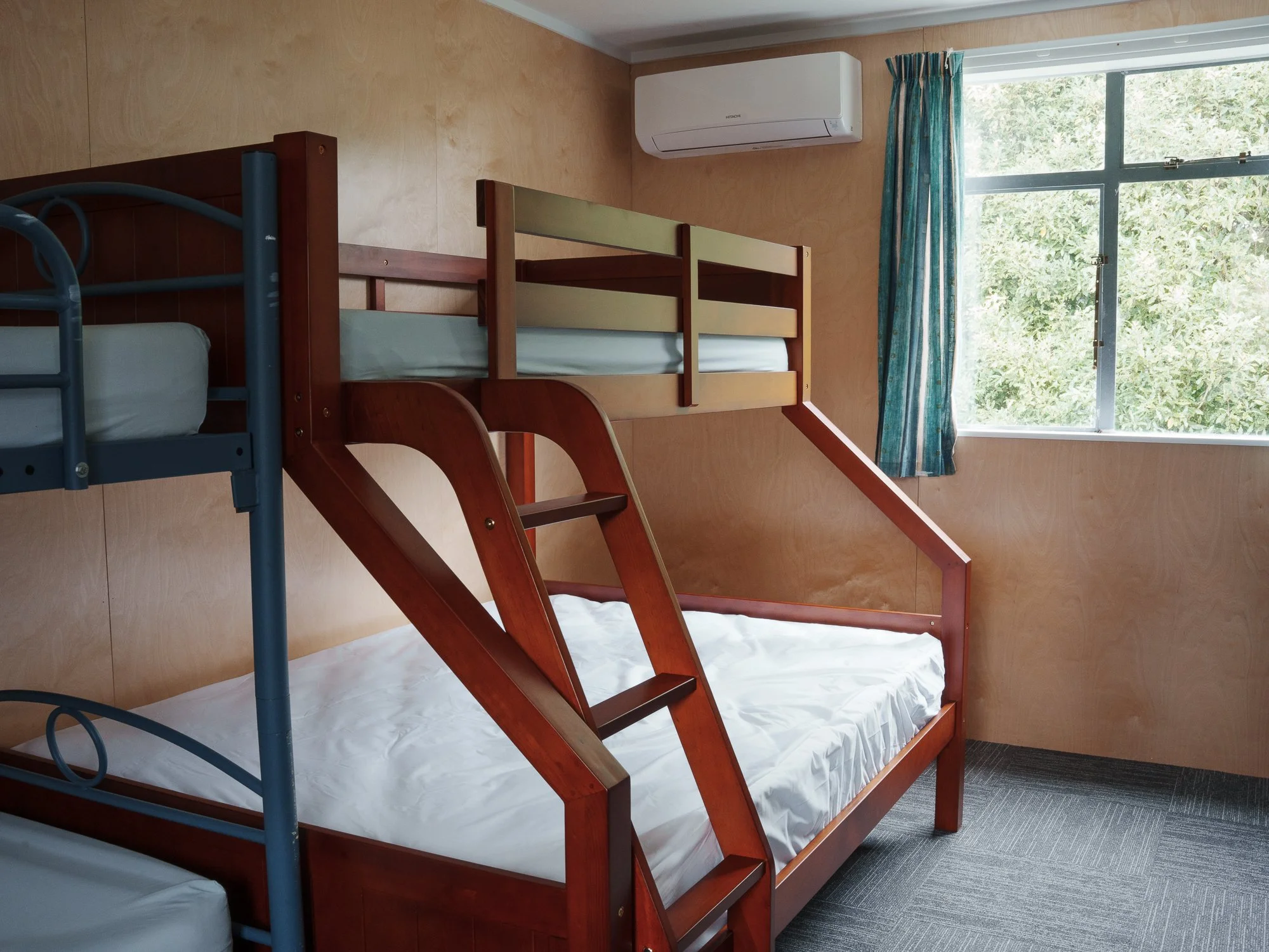 Room with a wooden bunk bed with a slide, a window with green trees outside, wood-paneled walls, an air conditioning unit, and a gray carpeted floor.