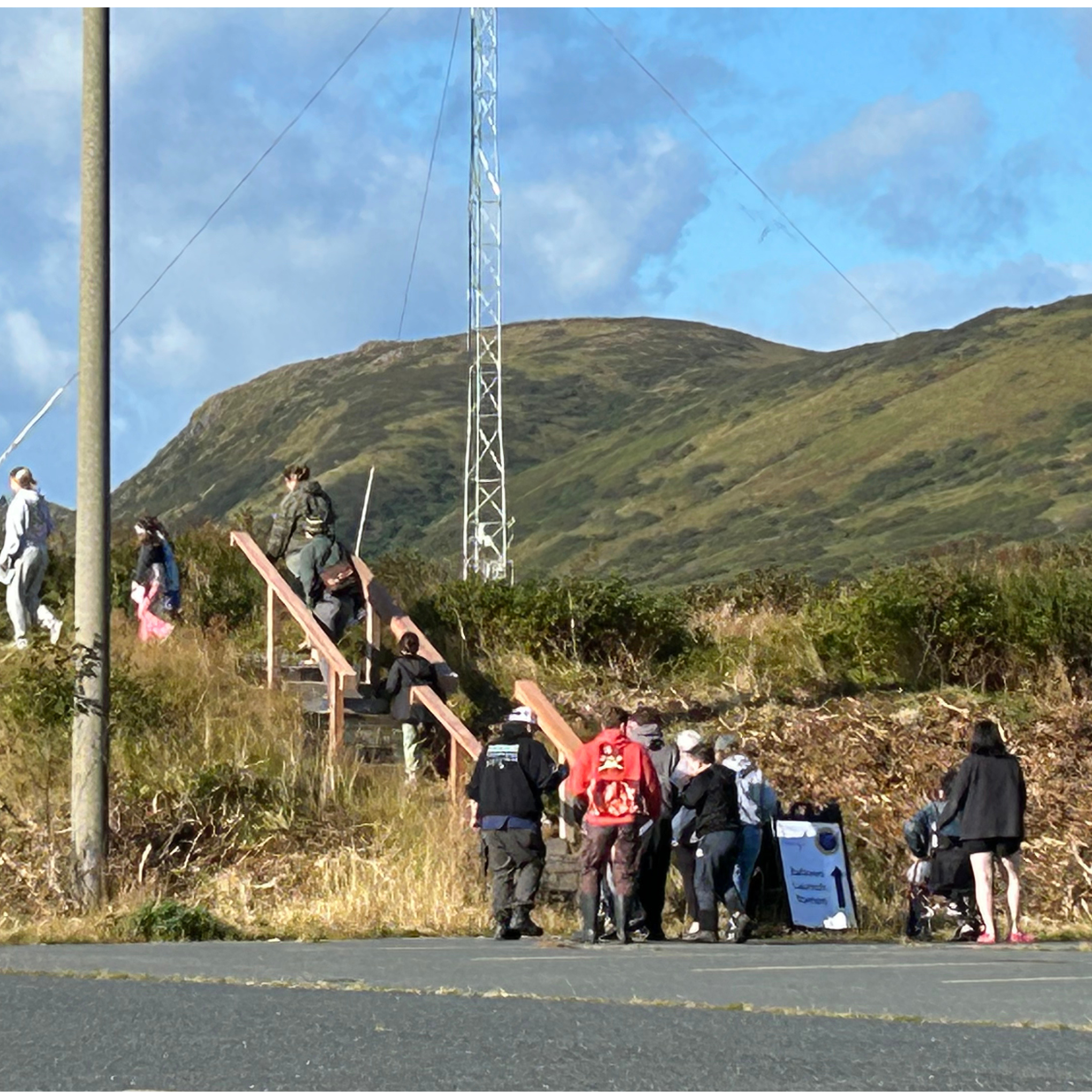 Group of people walking up outdoor stairs on a hillside near a mountain with a metal tower and cloudy sky in the background.