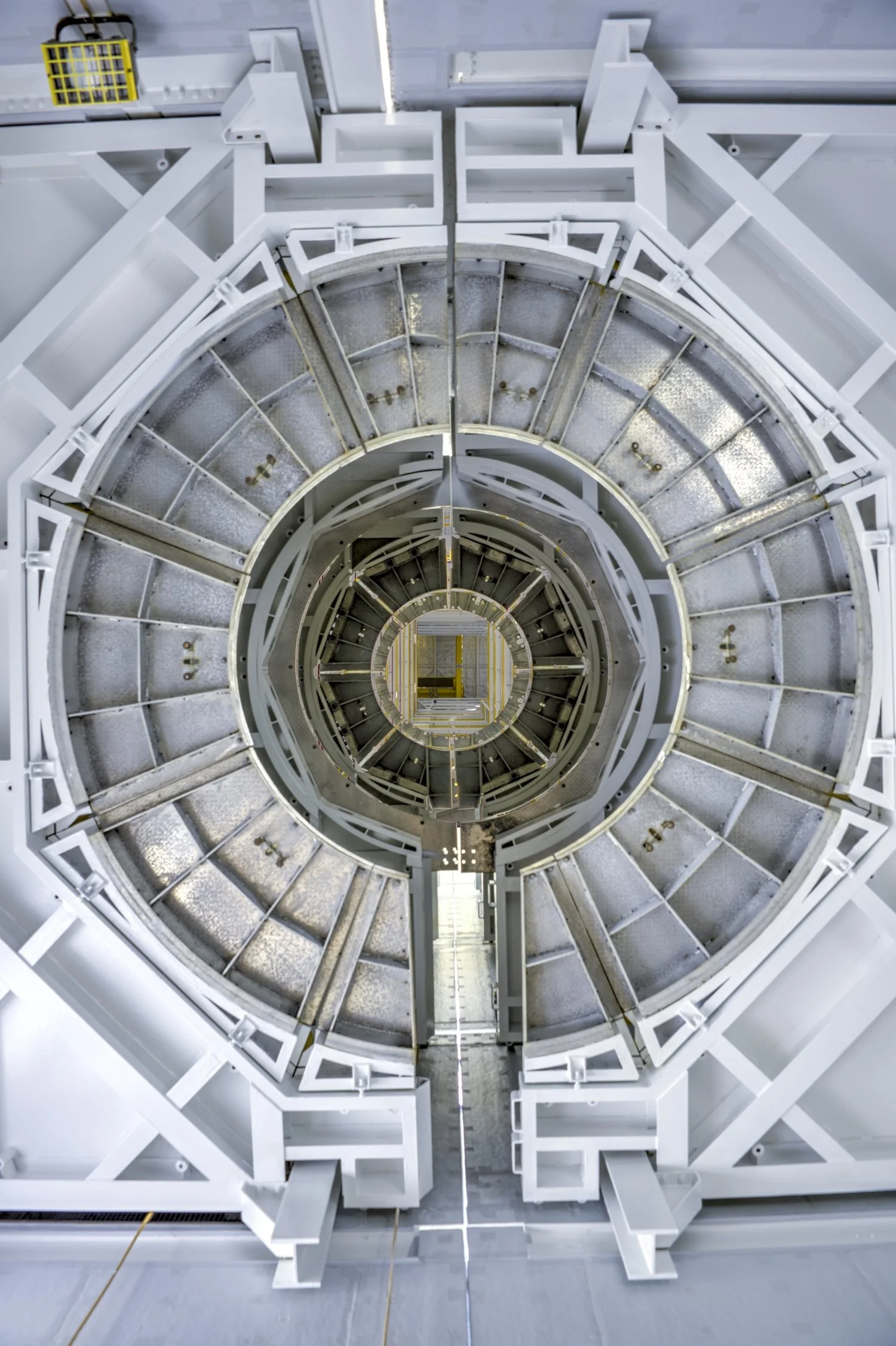 Looking down a large, multi-layered spiral staircase or tunnel made of metal and painted white, with visible structural supports and a ladder in the center.