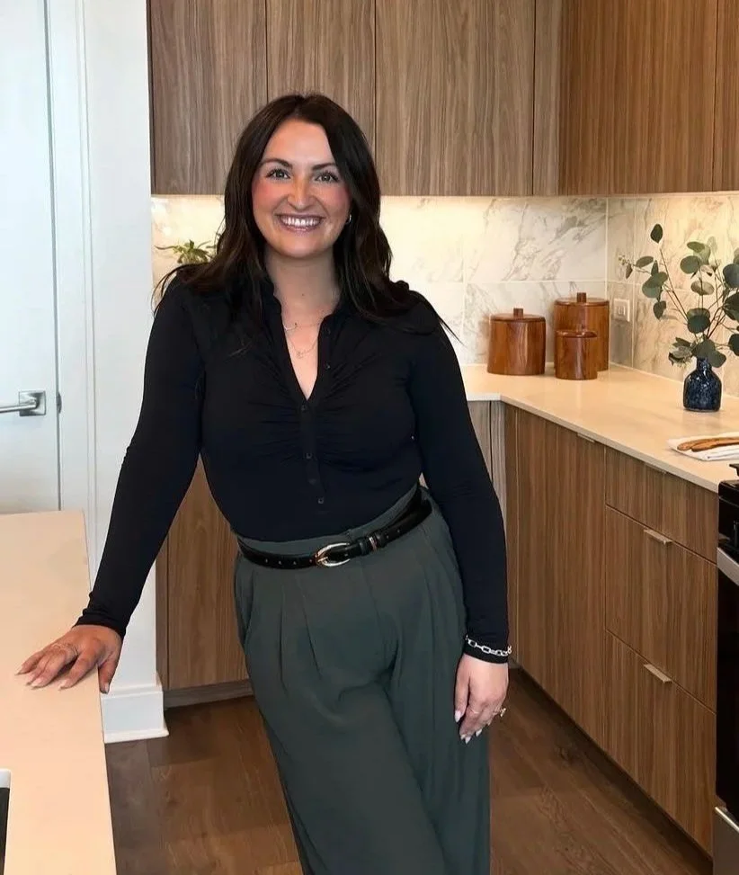 A girl smiling at the camera while standing in the kitchen, leaning against a kitchen island. She is wearing a black blouse with dark green gray trousers and a thin black belt.