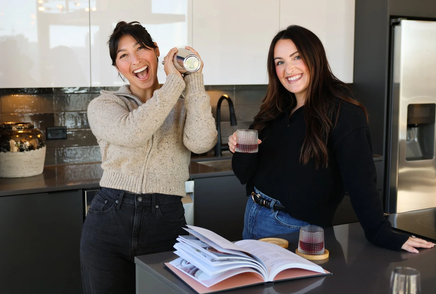 Two girls smiling at the camera, standing at a kitchen island. The girl on the left is shaking a barista shaker and is wearing a cream zip up jacket with black jeans. The girl on the right is wearing a black sweater and blue jeans, holding a drink.