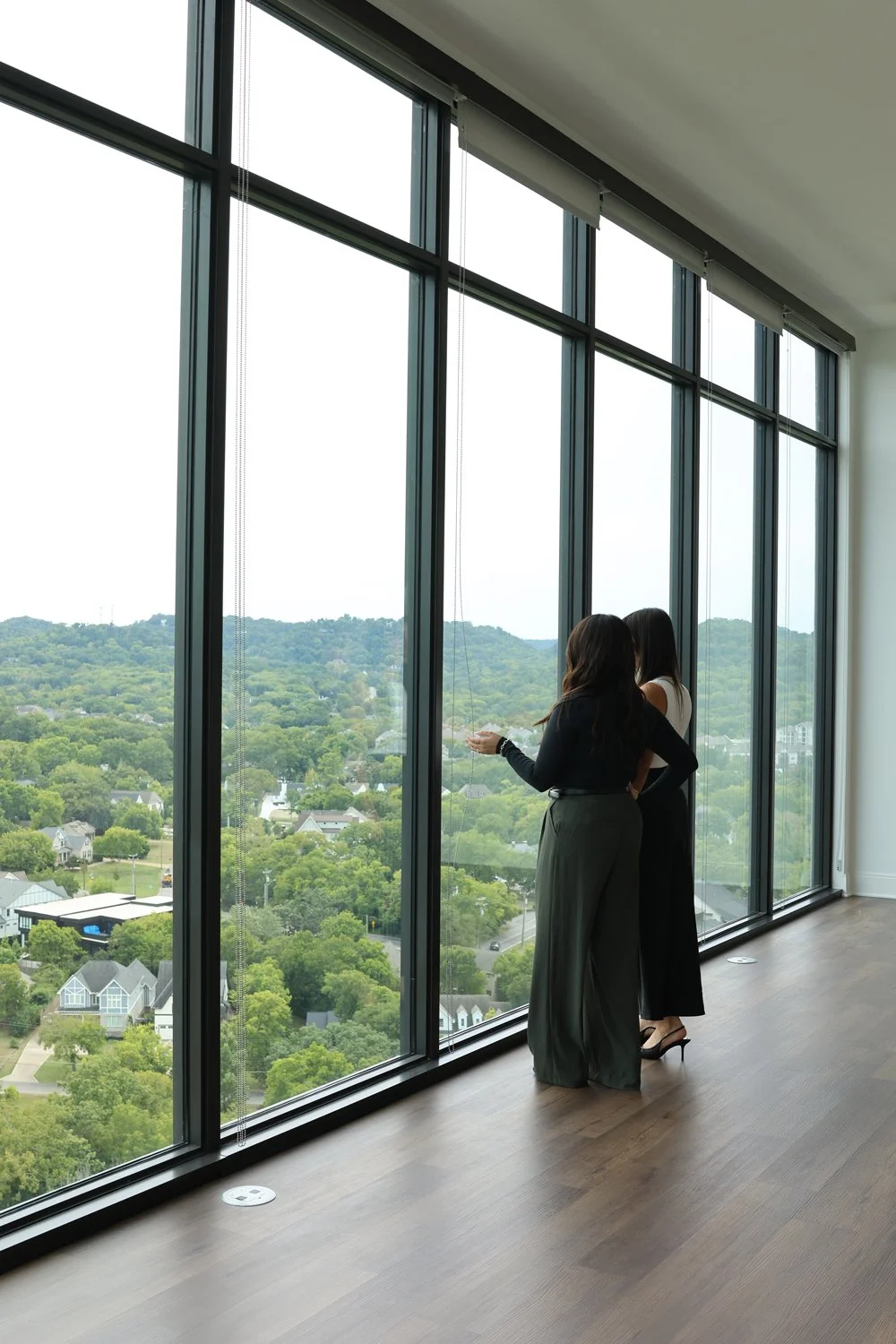 The backside of two girls looking out floor to ceiling windows at a neighborhood filled with trees.
