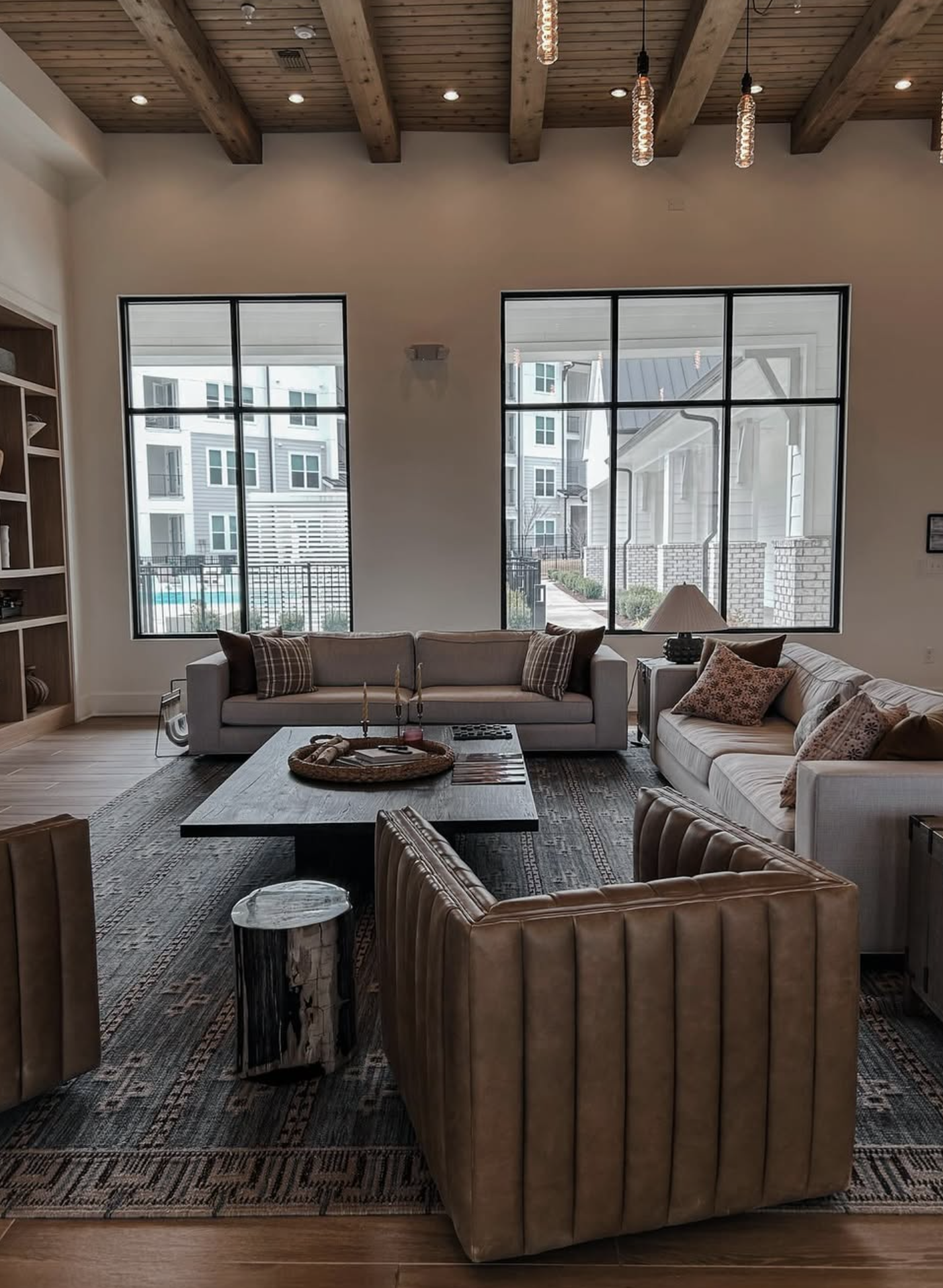 Living room of a home with big windows that show a gated pool and other condos. The living room has 2 dark beige couches, a dark wood coffee table, and a brown leather chair.