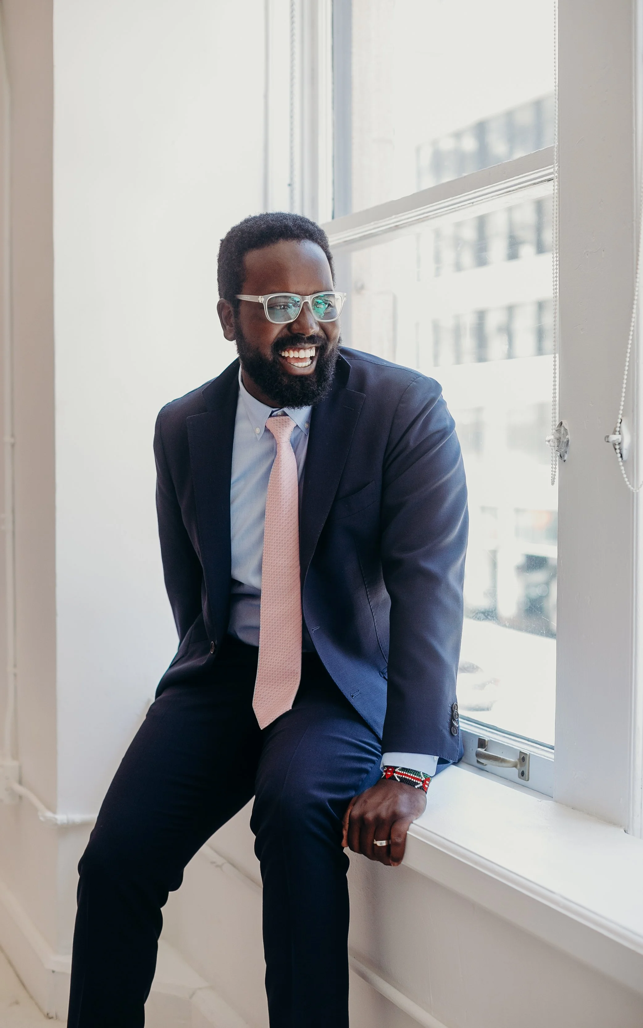 A smiling man in a business suit sitting by a window in a modern office building.