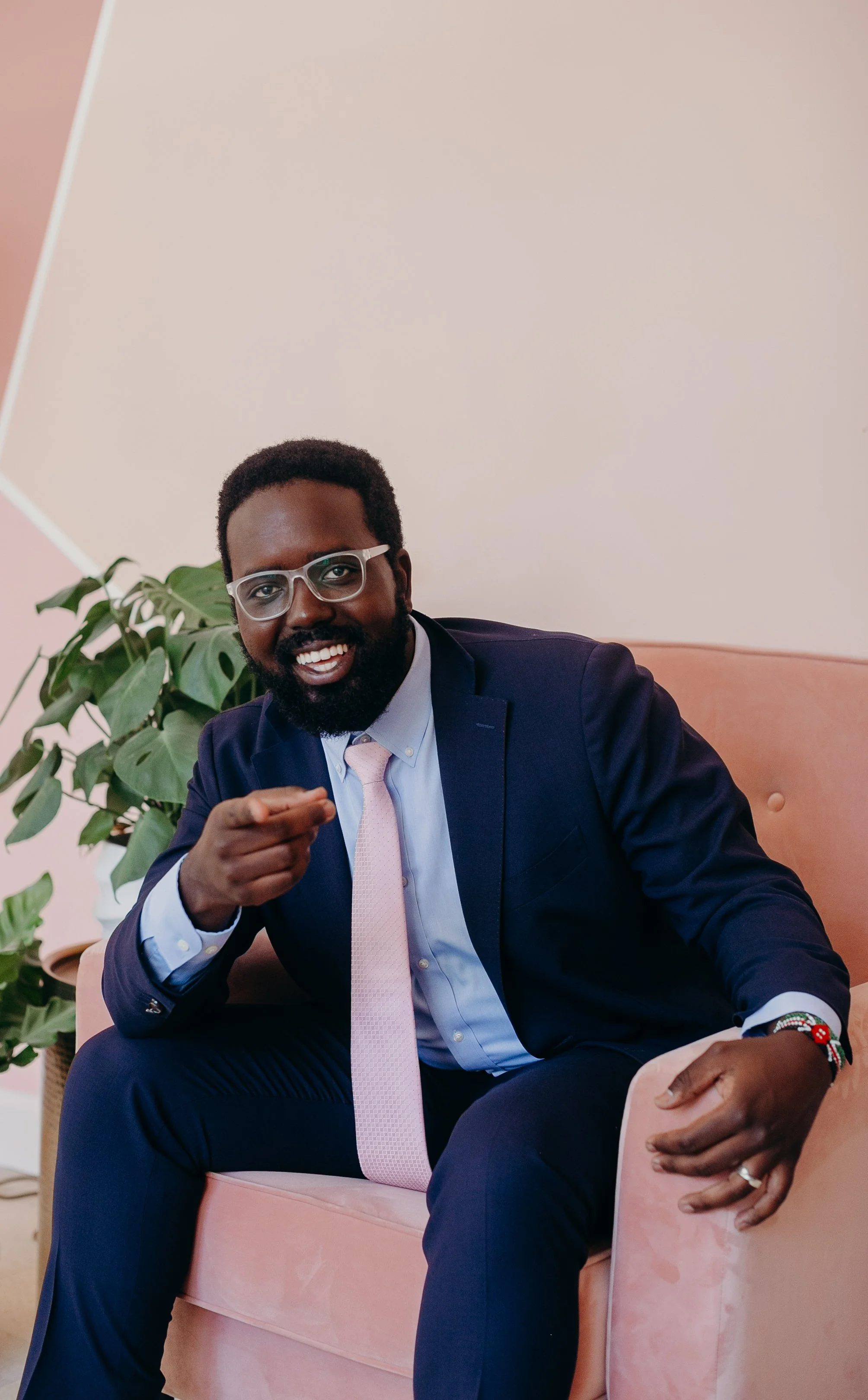 A smiling man in a dark blue suit, light pink tie, and glasses sitting on a pink sofa, pointing towards the camera, with a green plant in the background.