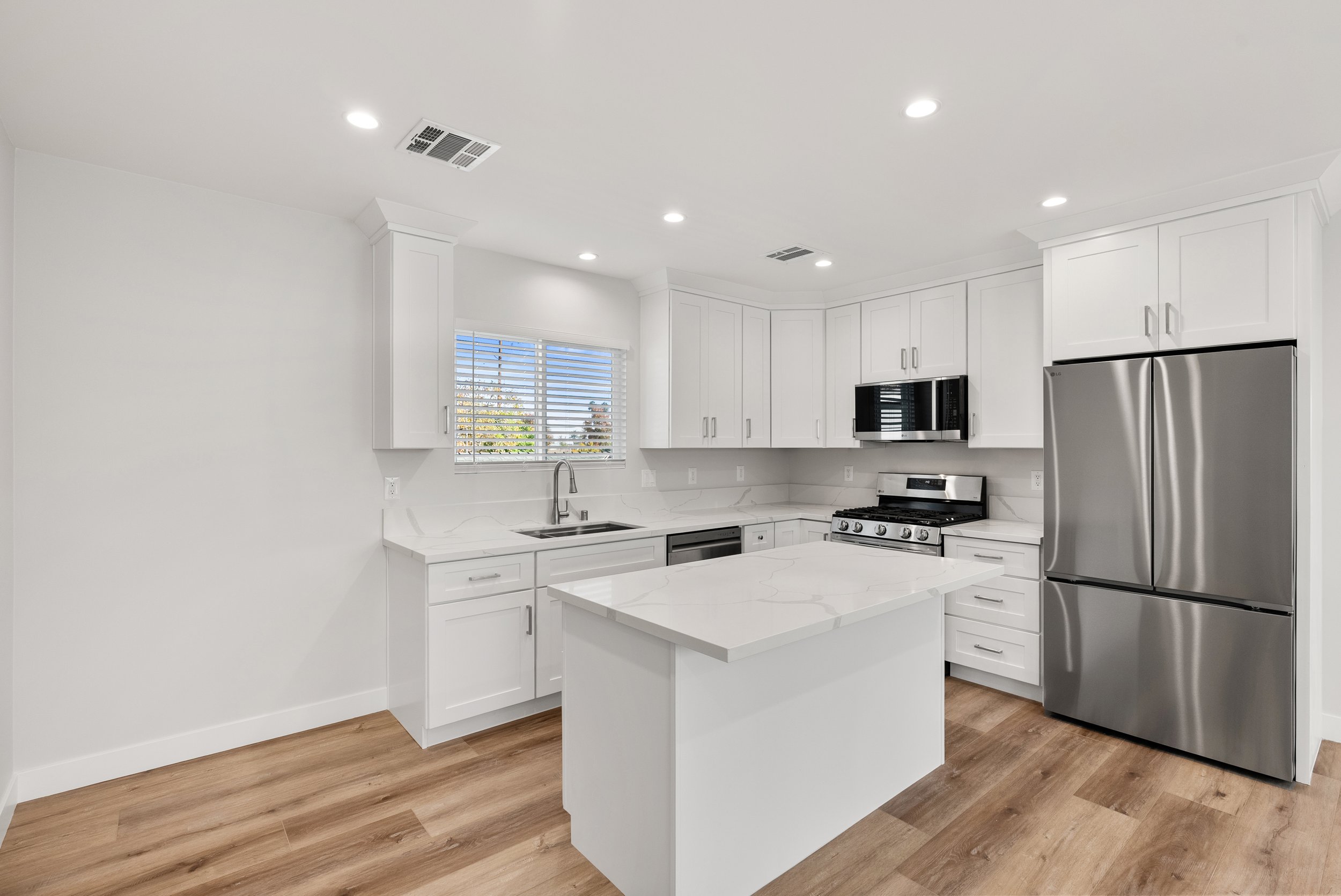 Modern kitchen with white cabinets, stainless steel appliances, marble countertops, hardwood flooring, and a window with blinds.