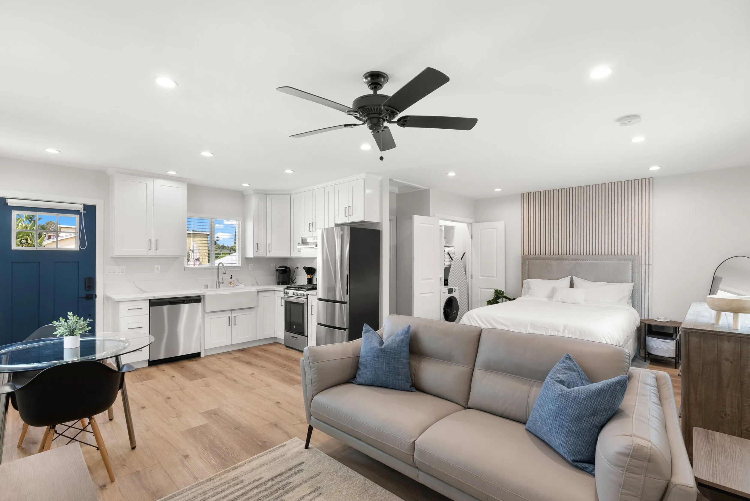 Open-plan studio apartment with kitchen, living area, and sleeping space. Kitchen has white cabinets, stainless steel appliances, and a small window. Living area features a beige sofa with blue pillows and a round glass dining table with black chairs