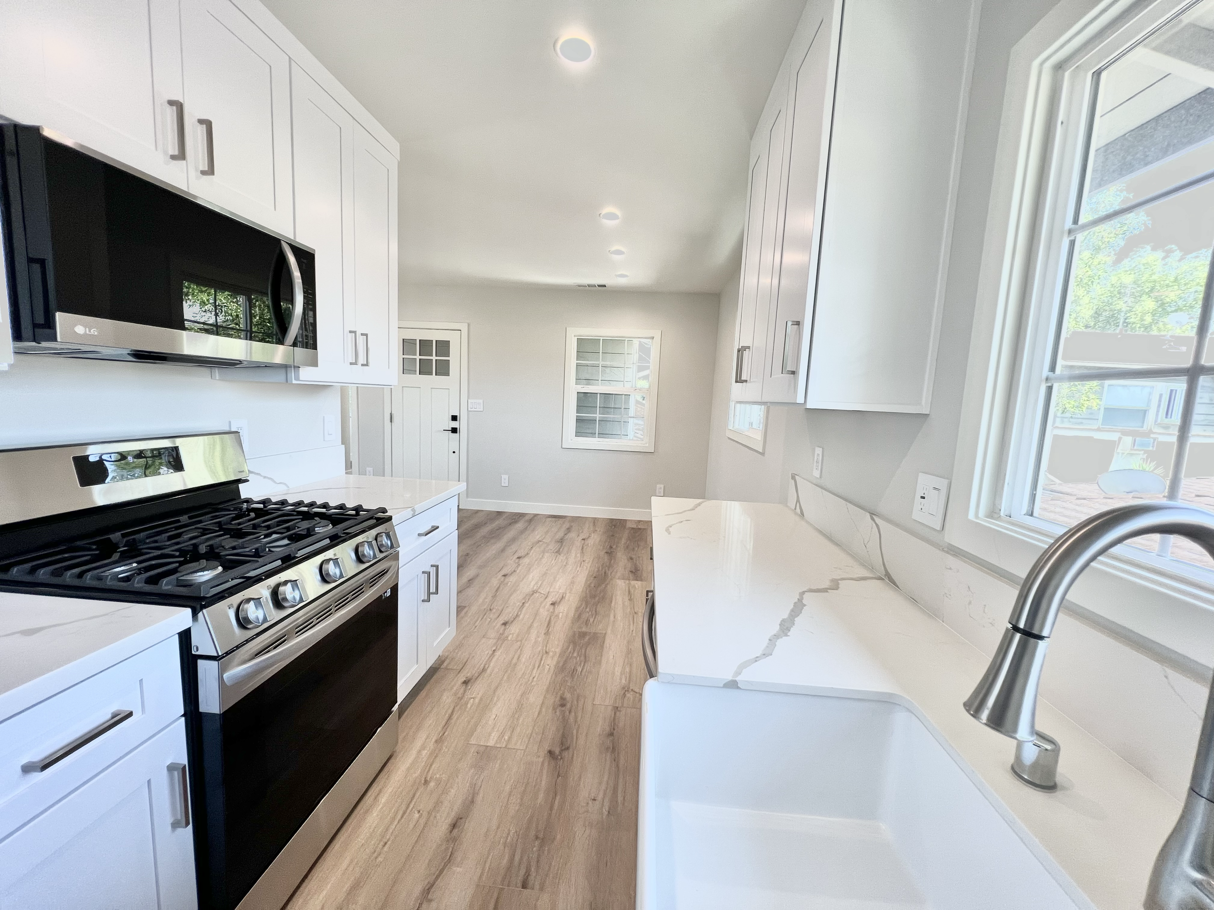 A modern kitchen with white cabinets, a marble countertop, a stainless steel oven and microwave, a large window, and a sink with a curved faucet.
