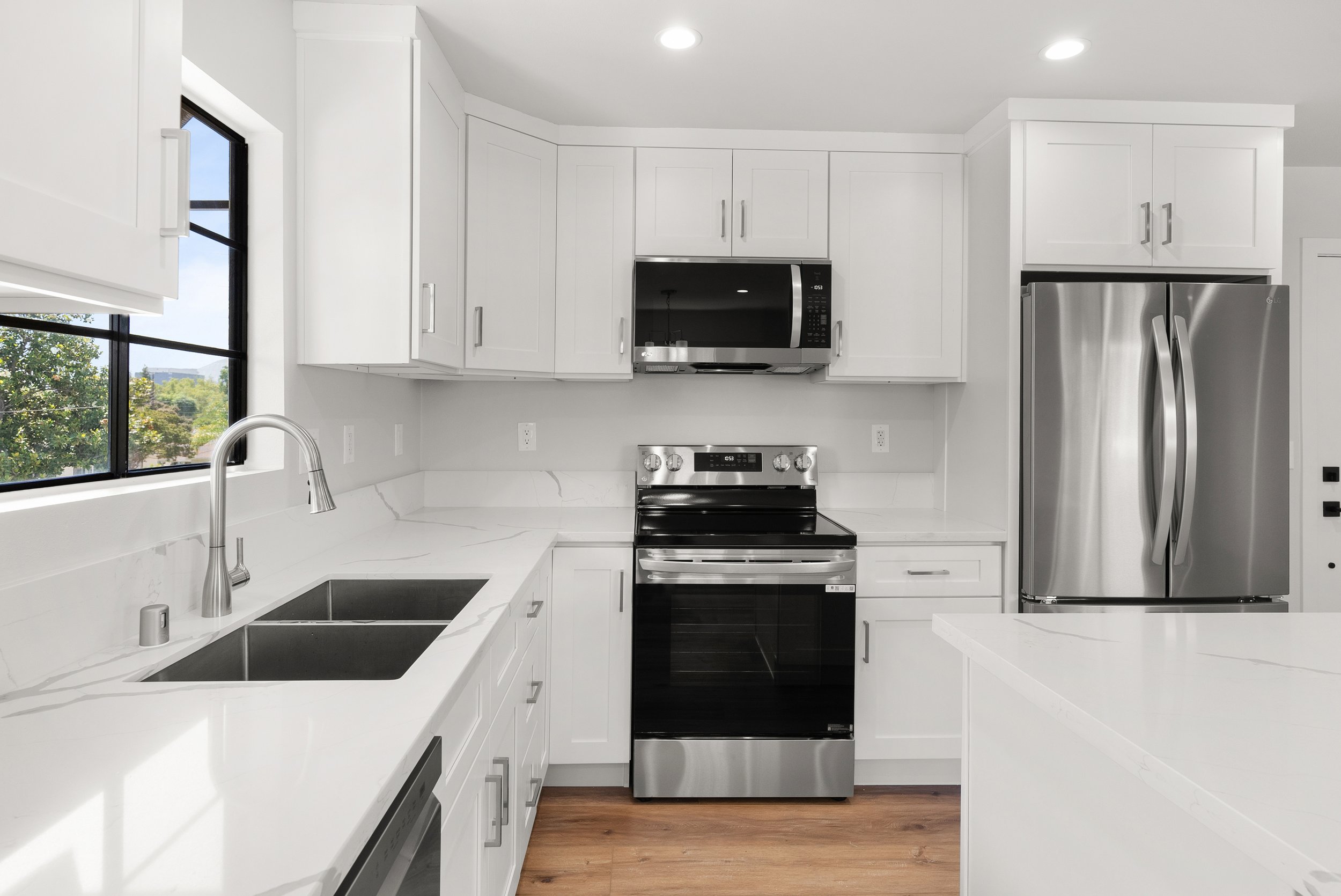 Modern white kitchen with stainless steel appliances, black microwave above black stove, white cabinets, and a window with a view of greenery.