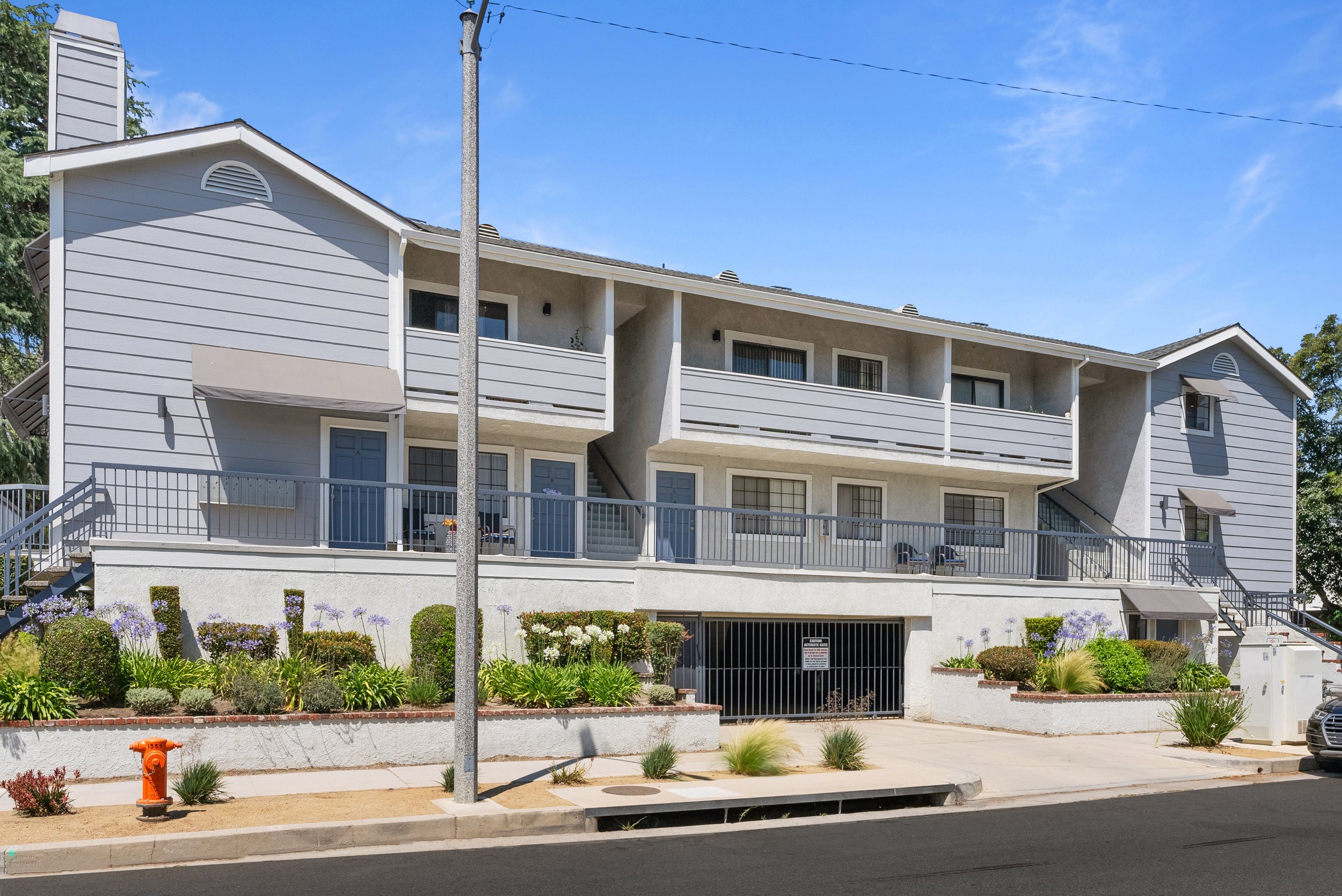 A residential apartment building painted in light gray, with multiple stairs leading to front doors, balconies, and a parking garage below. The building is surrounded by a landscaped garden with various flowers, bushes, and plants, and has a fire hydrant and utility boxes in the foreground.